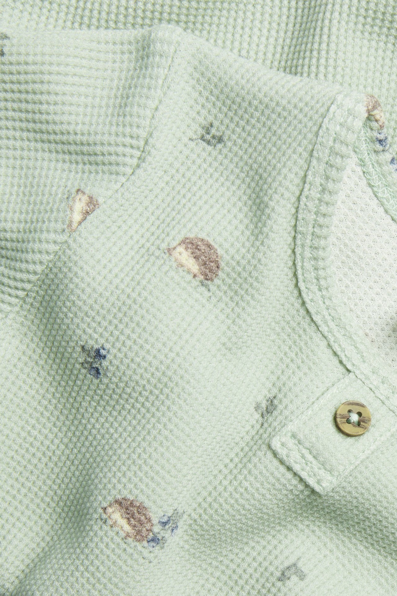 Zbliżenie zielonego, długorękawowego body waflowego z wzorem jeży i ozdobnym drewnianym guzikiem.