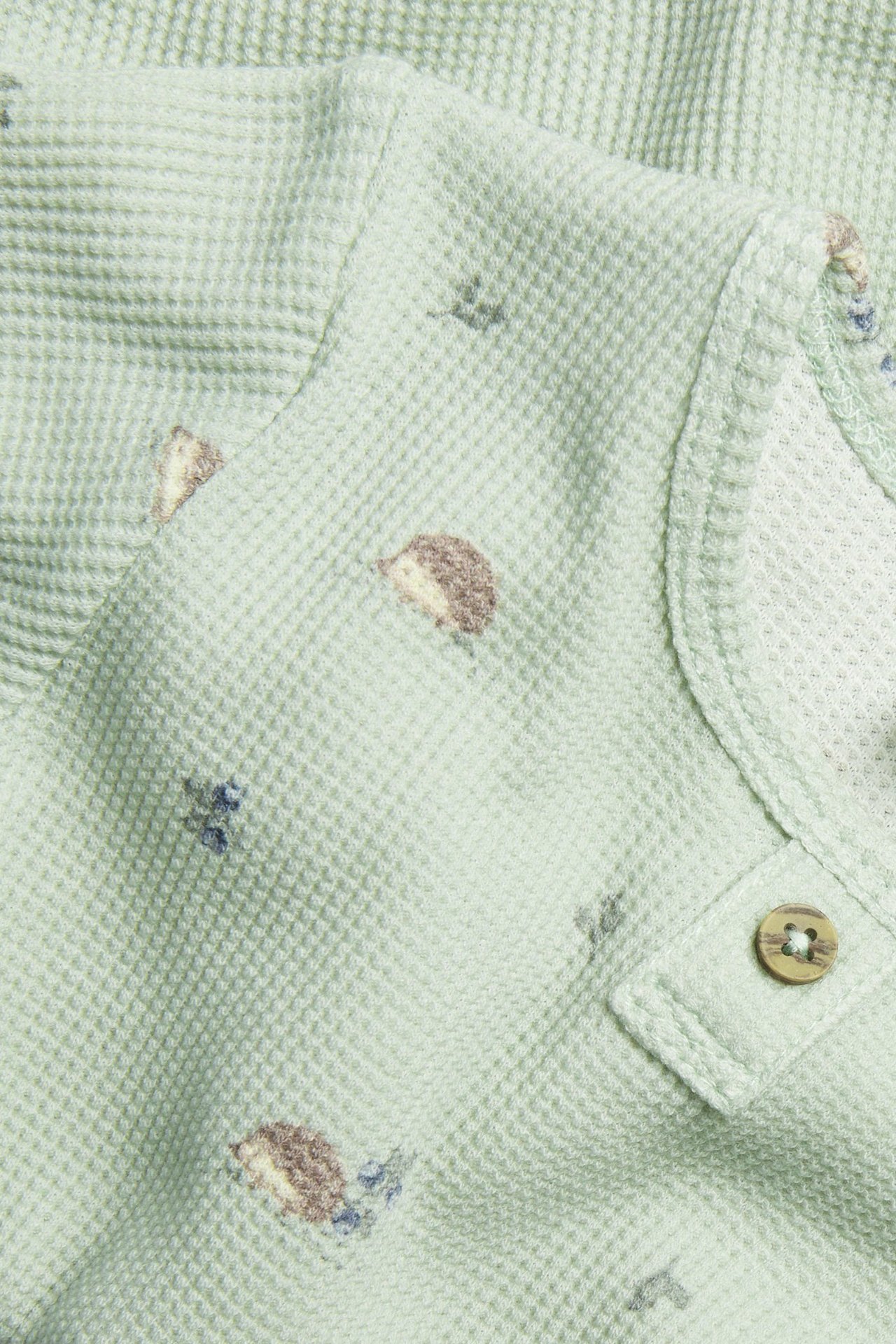 Närbild på en grön långärmad body i våffelkvalitet med igelkottsmönster och en dekorativ träknapp.