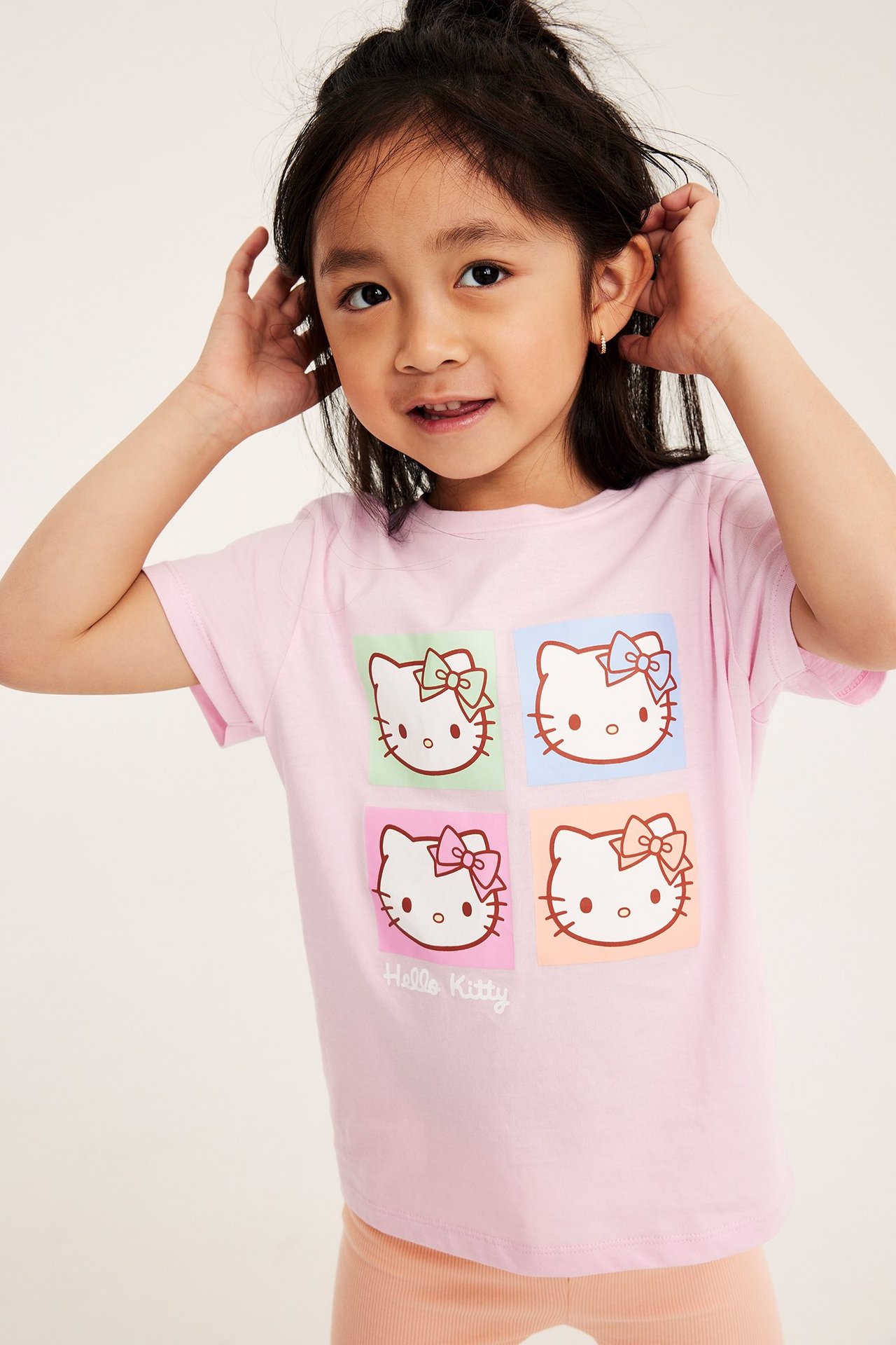 Framifrån, ett barn i en rosa T-shirt i bomull med Hello Kitty-tryck, över ljusorange ribbade shorts.