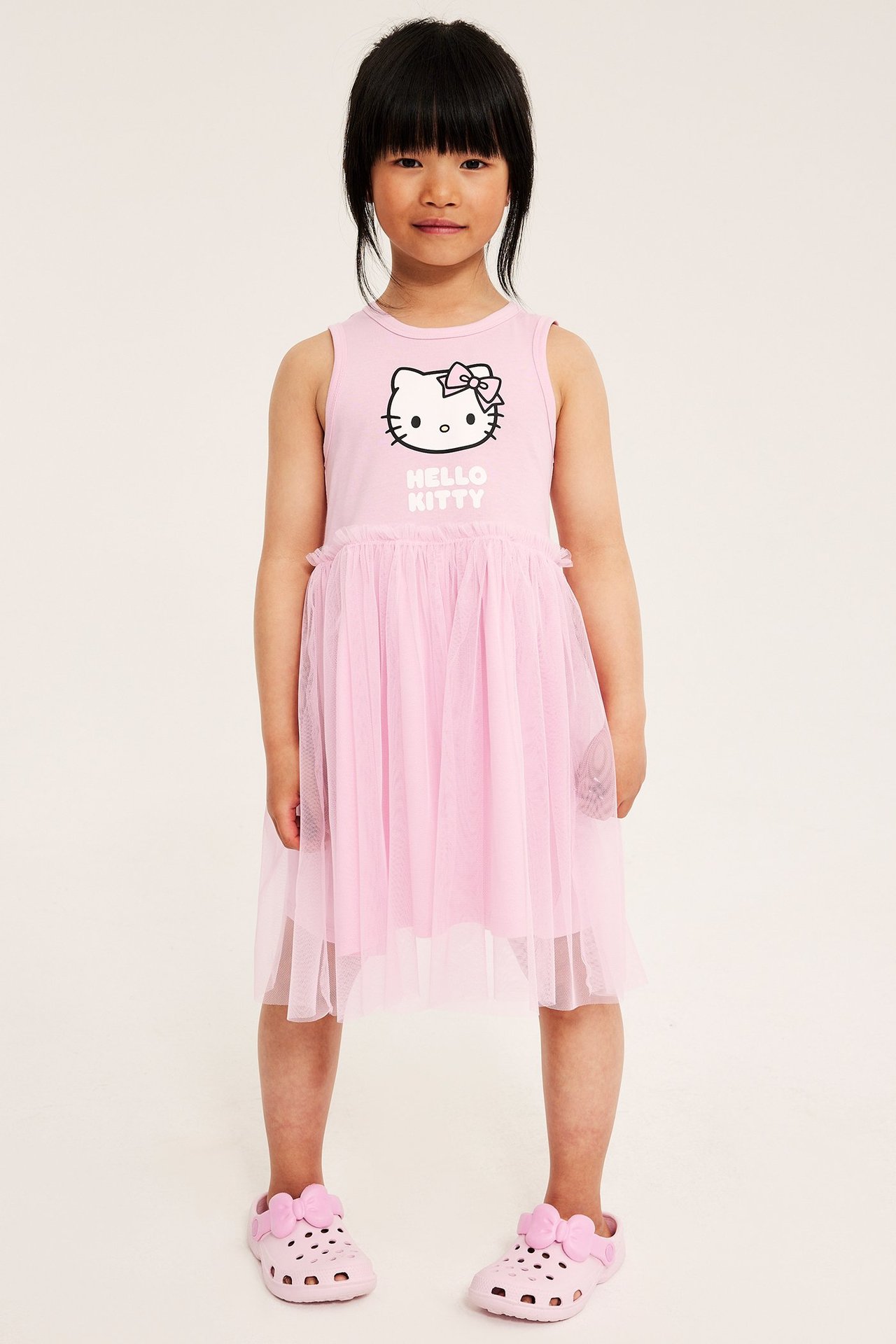 Frontbilde av et barn i en rosa ermeløs Hello Kitty jerseykjole med tyllskjørt og rosa crocs.
