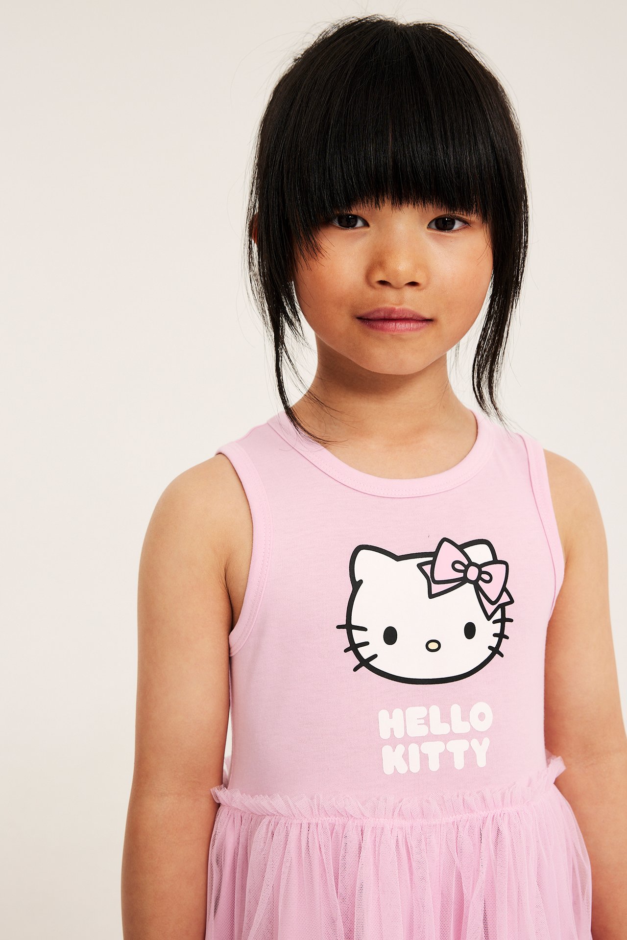 Lähikuva edestä lapsesta vaaleanpunaisessa hihattomassa Hello Kitty -jersey mekossa, jossa on rypytetty tyllihame.