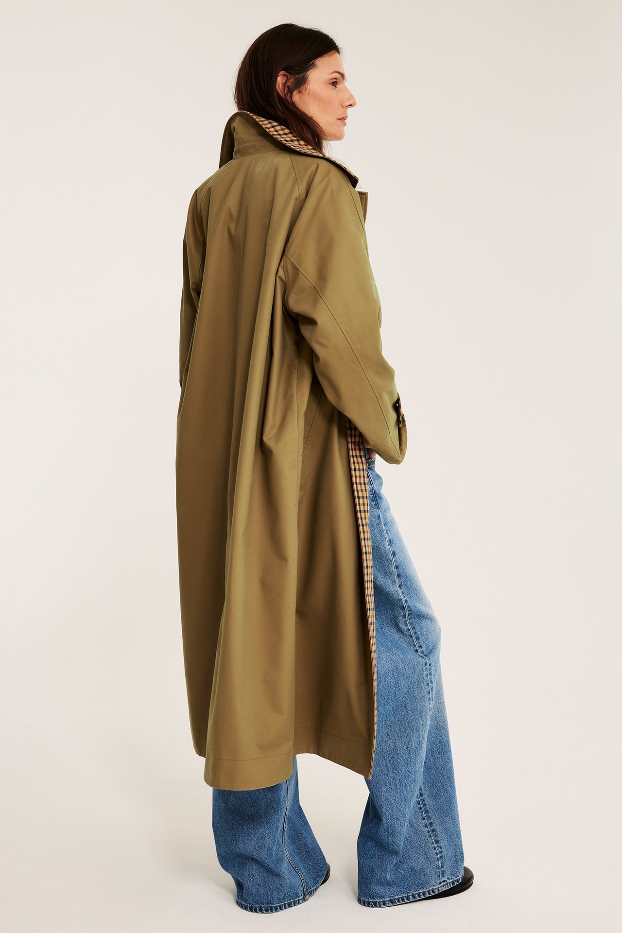 Sideprofil av en kvinne i en lang, løs beige vendbar kåpe med rutete fôr, over vide blå jeans.