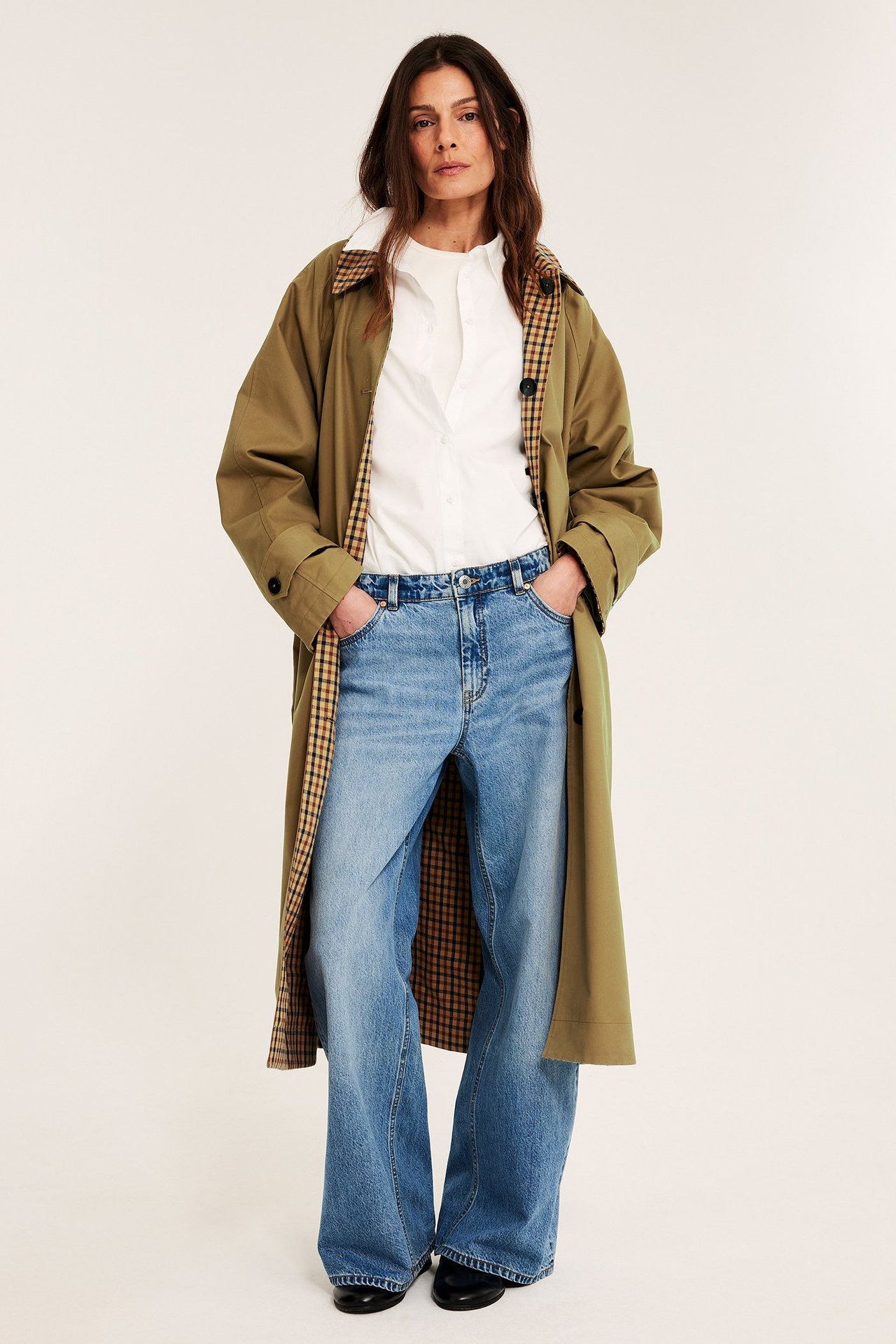 Framifrån: Kvinna bär öppen beige vändbar kappa med rutigt foder, vit skjorta och vida ljusblå jeans.