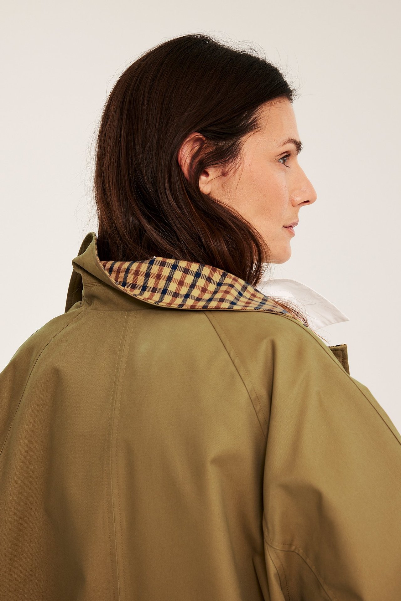 Sidebilde av kvinne i beige vendbar kåpe, med rutete fôr synlig ved krage over hvit skjorte.