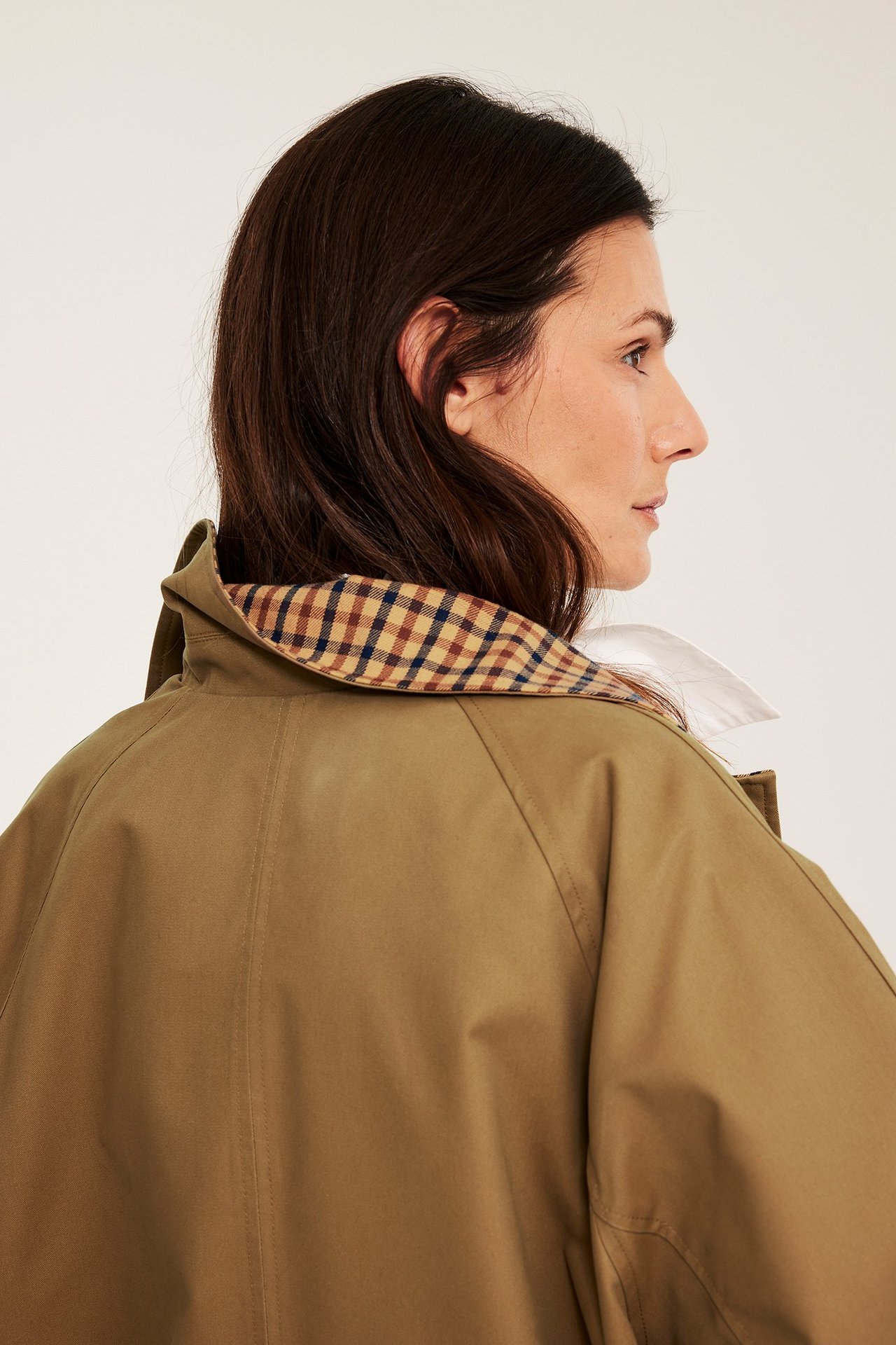 Widok z boku kobiety w beżowym dwustronnym trenczu z kratkowanym kołnierzem na białej koszuli.