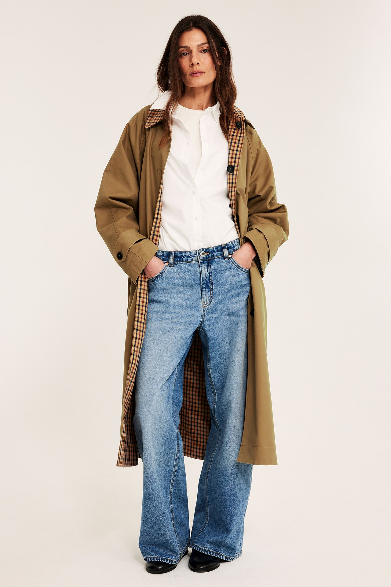 Framifrån: Kvinna i beige vändbar trenchcoat, vit skjorta, ljusblå jeans och svarta skor.