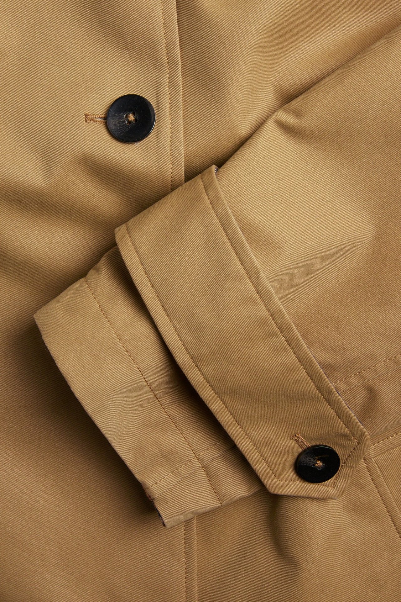 Zbliżenie beżowego, dwustronnego trenczu, ukazujące czarne guziki na plisie i mankiecie rękawa.
