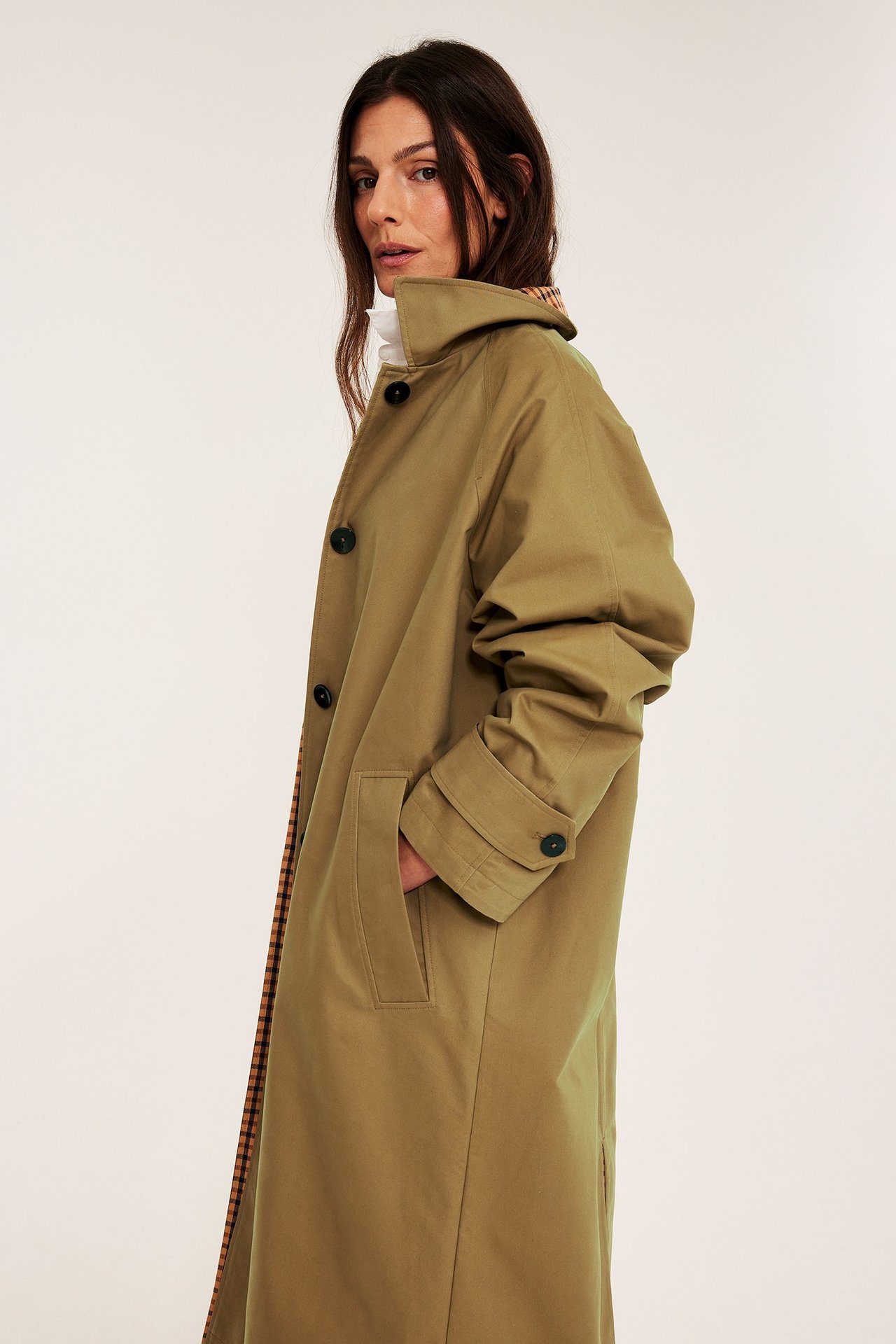 Sidovy av kvinna i beige vändbar trenchcoat med rutigt foder, mörka knappar, handen i fickan.
