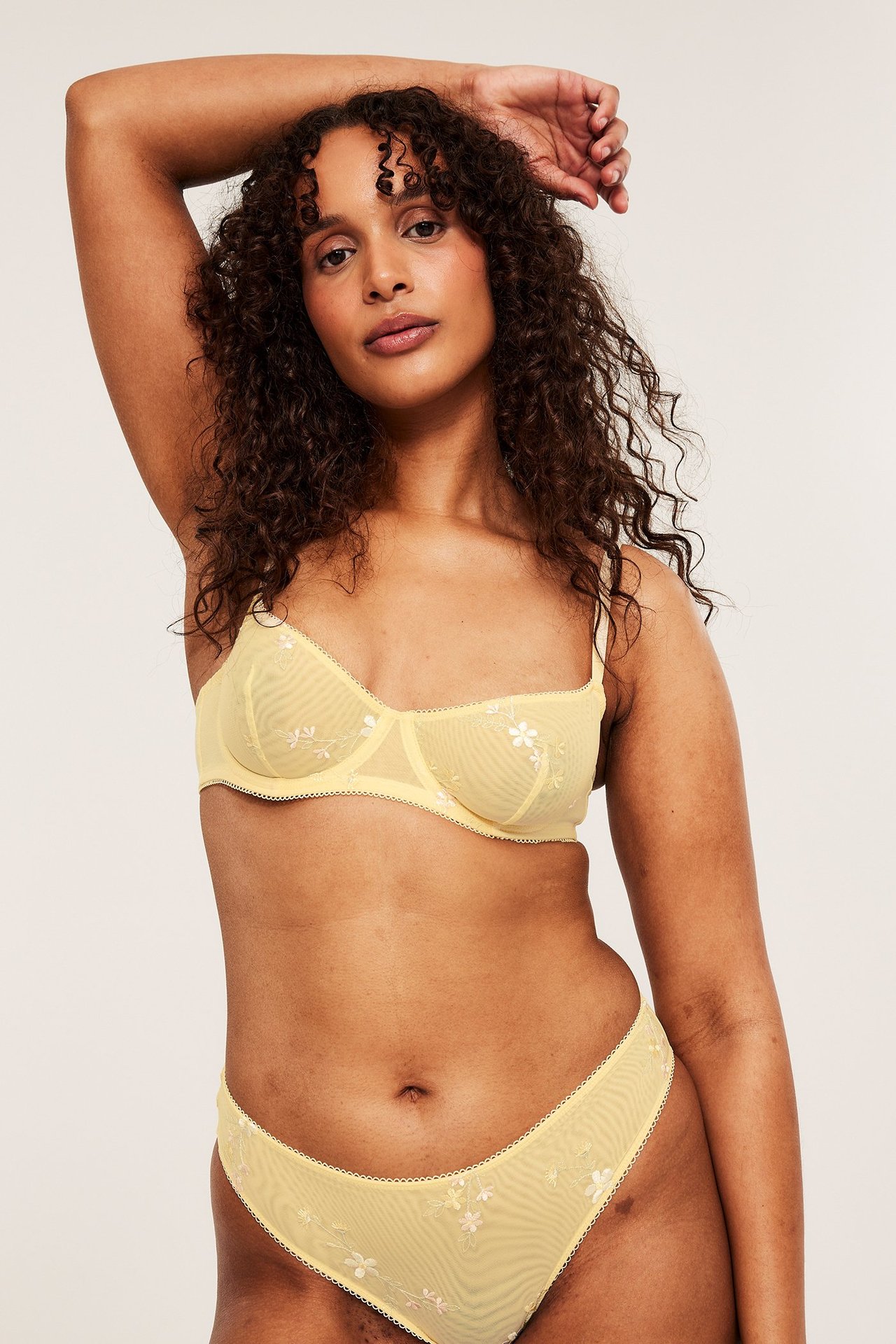 Forfra: Kvinne i lys gul bøyle-BH i mesh med blomsterbroderi og matchende truse.