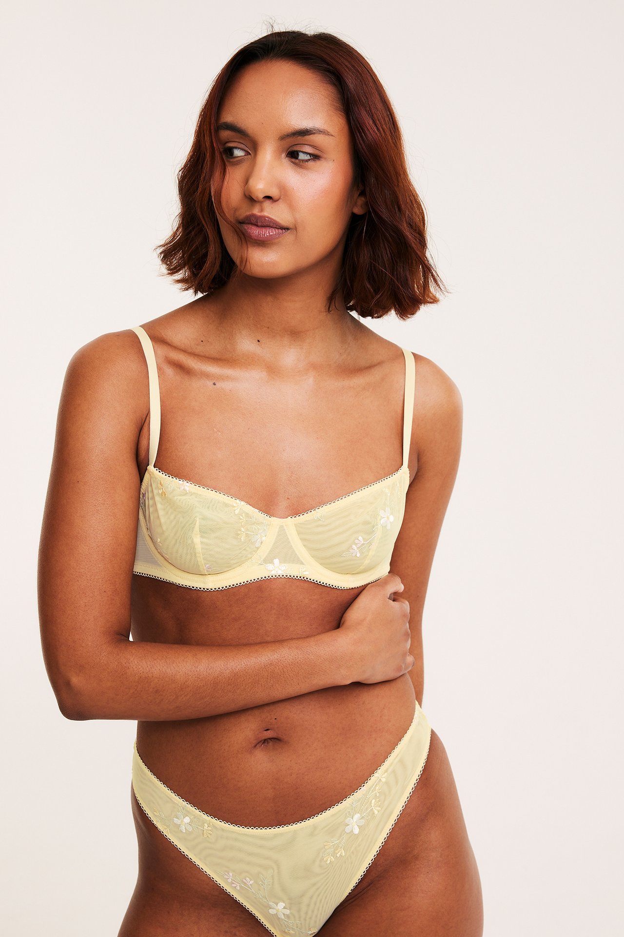 Frontvy: kvinna i ljusgul bygel-bh i mesh med vit blombrodyr och matchande trosor.