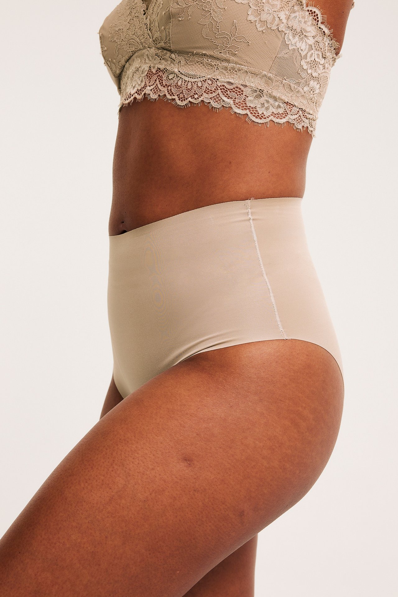 Kobieta z boku w beżowych, bezszwowych figach high-waist i jasnym koronkowym biustonoszu.