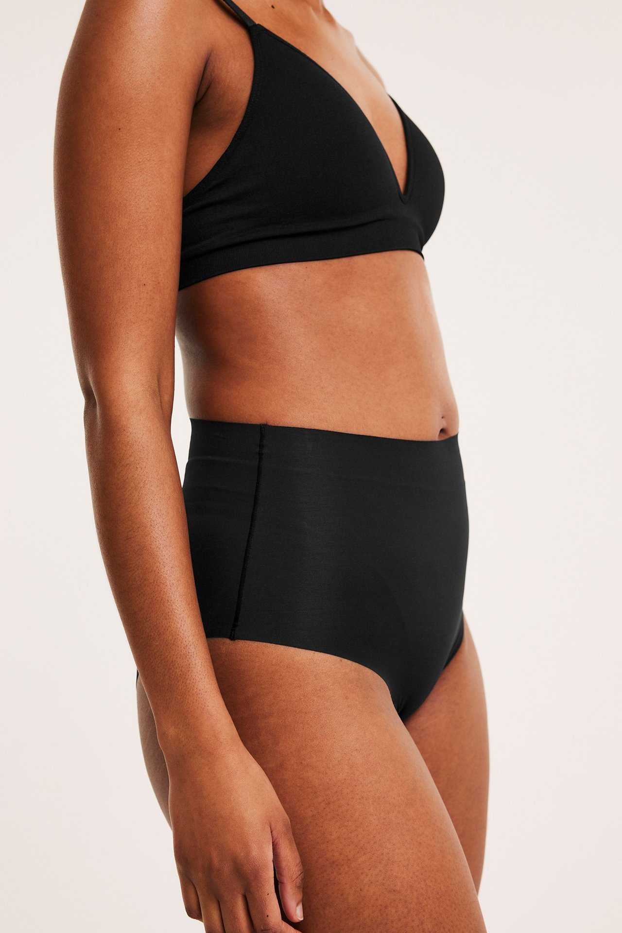 Sidebilde av kvinne i svarte sømløse høytlivs lett formende truser og matchende bralette.