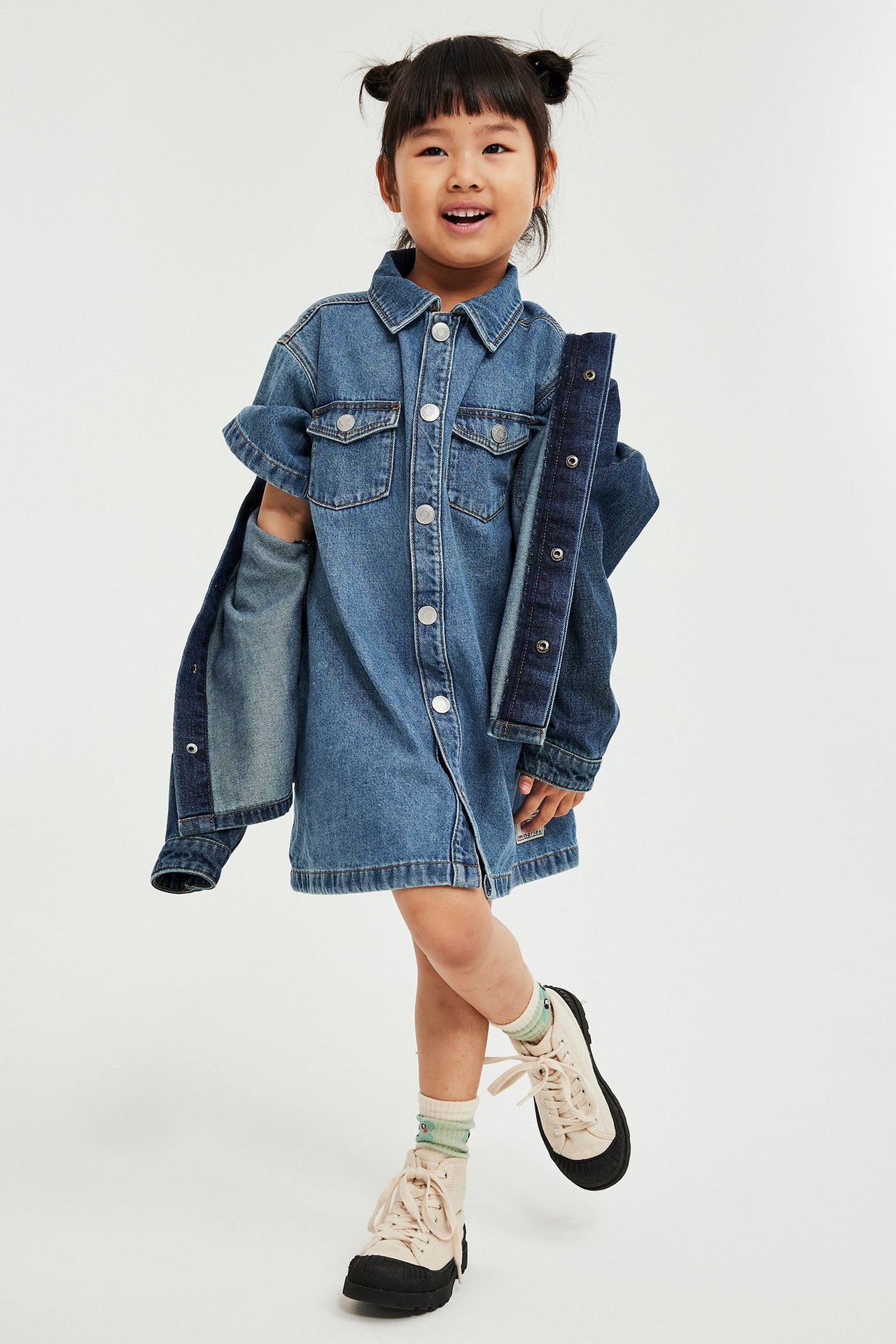 Dziecko w dżinsowej sukience z kołnierzem, guzikami, kieszeniami, jasnymi skarpetkami i beżowymi butami, widok z przodu.