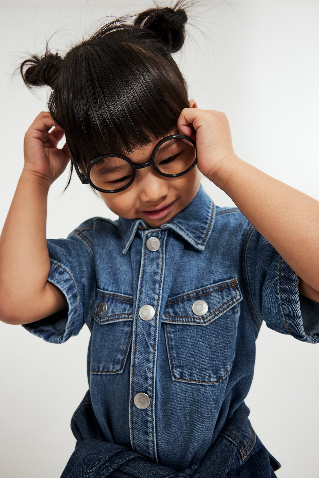 Zbliżenie na dziecko w dżinsowej sukience z zatrzaskami i czarnymi okrągłymi okularami.