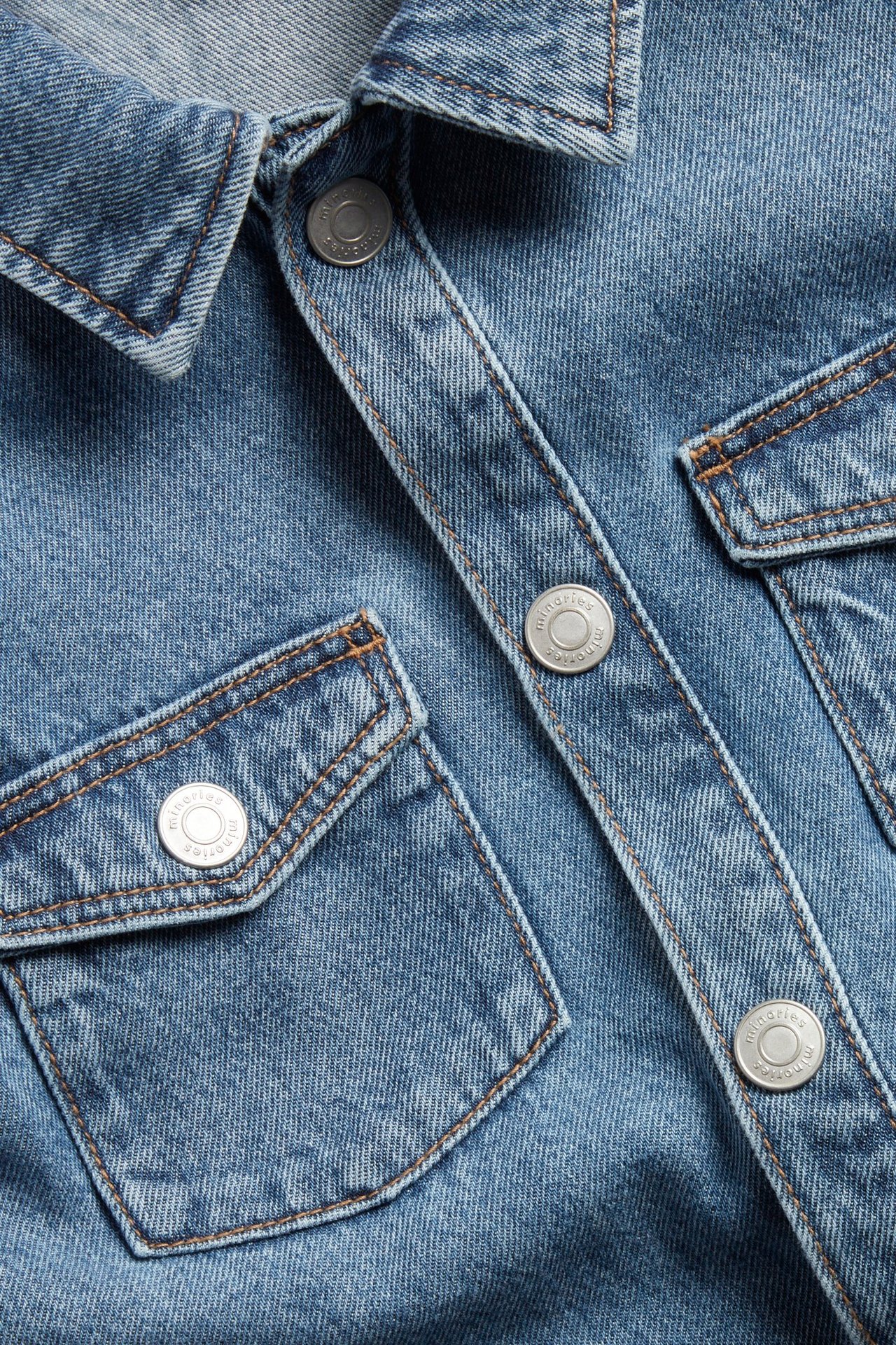 Zbliżenie jasnoniebieskiej sukienki dżinsowej z kołnierzem, zatrzaskami i kieszeniami na piersi z brązowymi szwami.