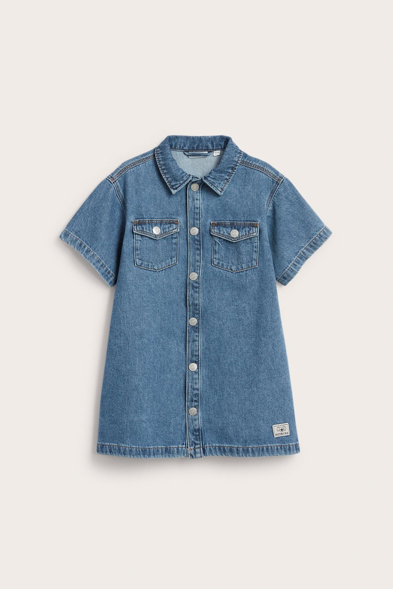 Widok z przodu: krótka niebieska sukienka jeansowa dla dziecka z kołnierzykiem, zatrzaskami i kieszeniami na piersi.