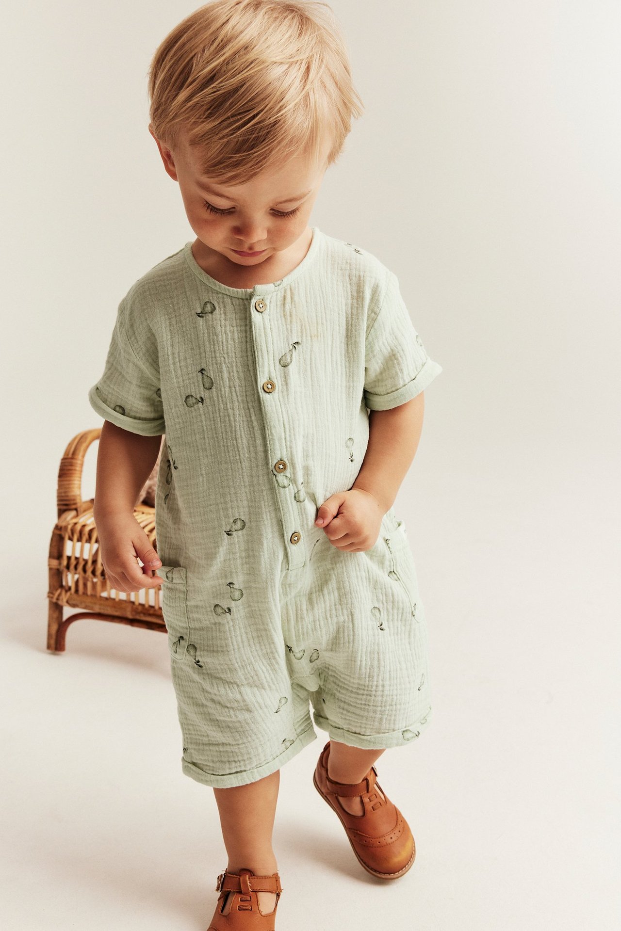 Frontvy, baby i ljusgrön päronmönstrad bomullsromper med knappar, fickor och bruna skor.