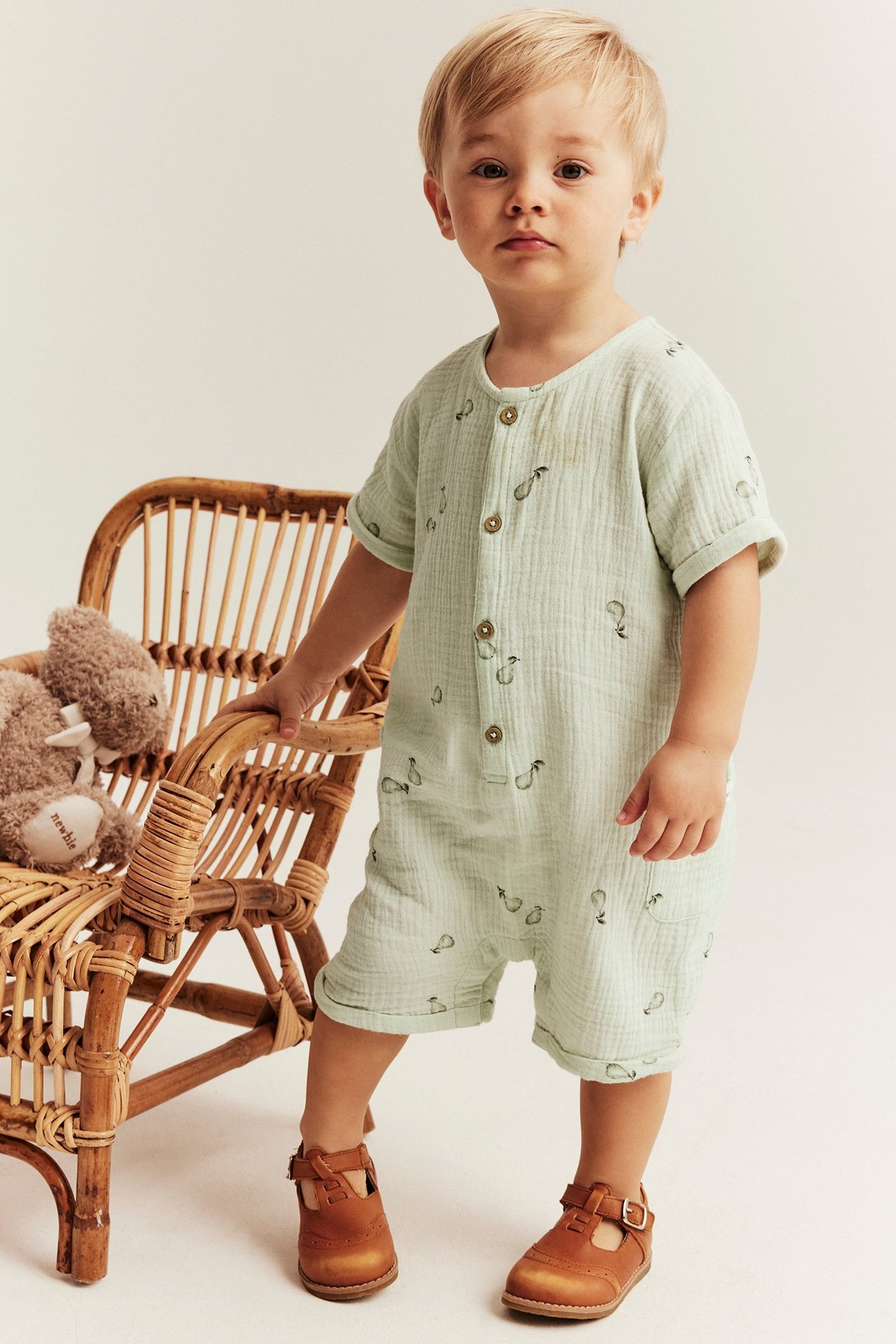 Frontvy av ett barn i ljusgrön vävd bomullsromper med pärnmönster, knappar fram och bruna T-remsskor.