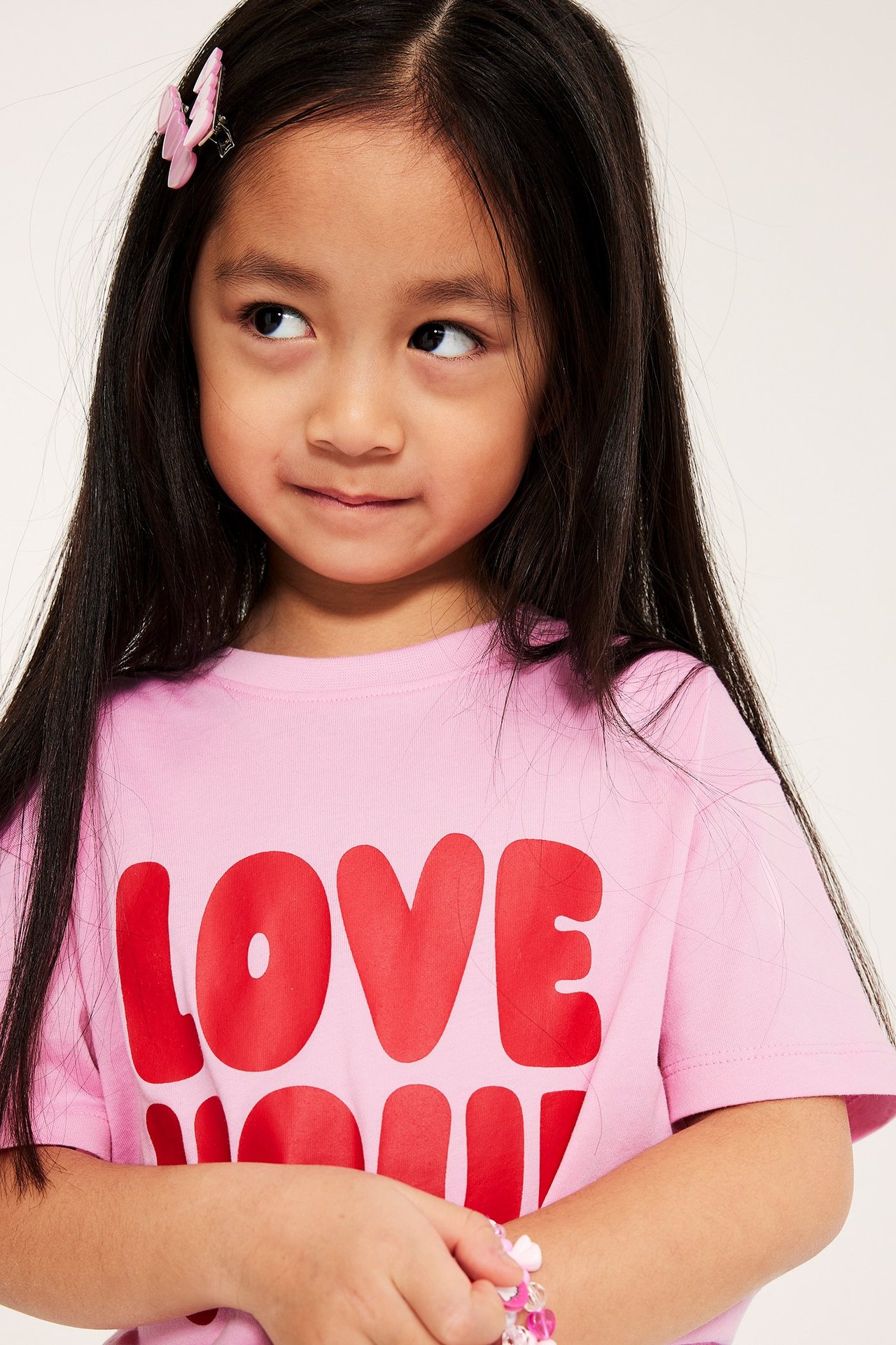 Närbild på ett barn som bär en ljusrosa T-shirt i bomull med rött texttryck, rosa fjärilshårklämma och pärlarmband.