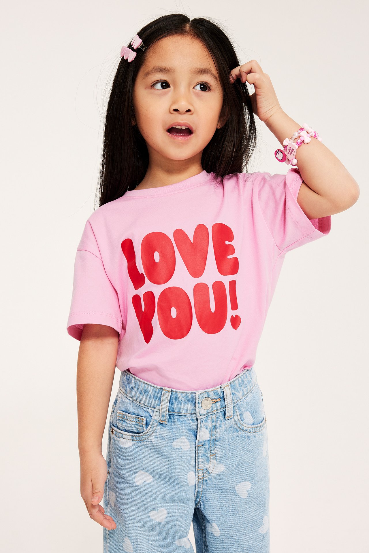 Dziecko nosi różowy T-shirt "LOVE YOU!", jasnoniebieskie dżinsy w serca, różowe spinki z kokardkami i różową bransoletkę.