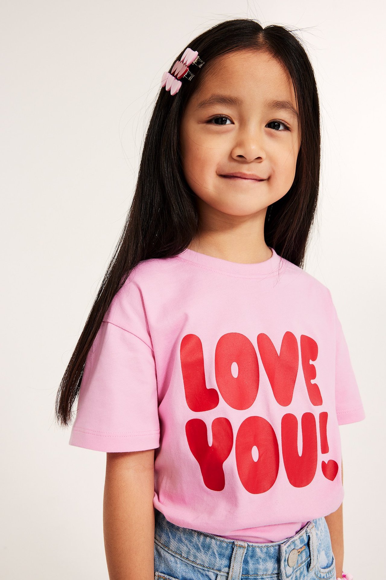 Dziecko w jasnoróżowej bawełnianej koszulce z czerwonym napisem 'LOVE YOU!', niebieskich dżinsach i różowych spinkach.