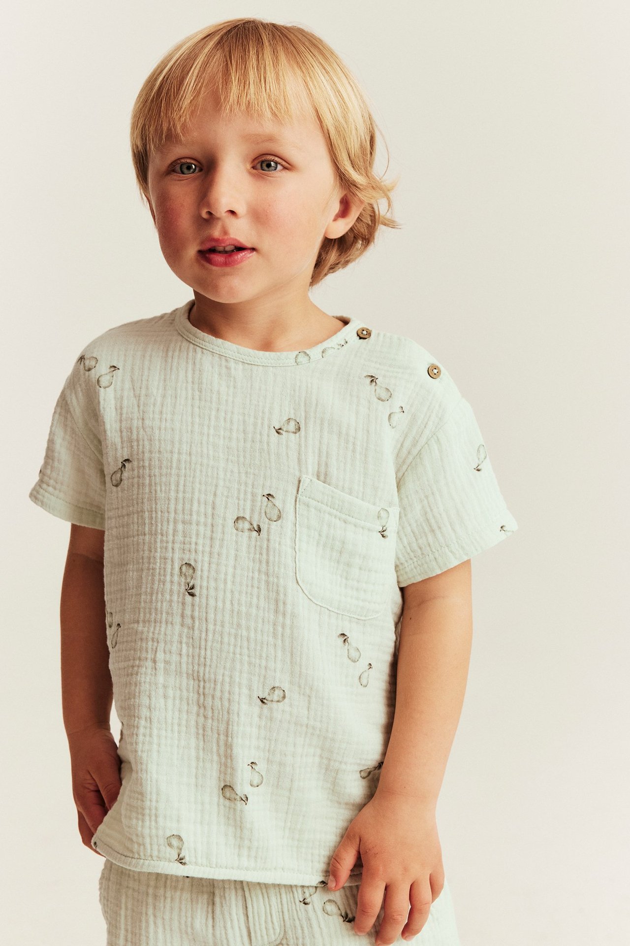 Lapsi yllään vaaleanvihreä päärynäkuvioinen musliinitoppi olkapäänapeilla ja rintataskulla.