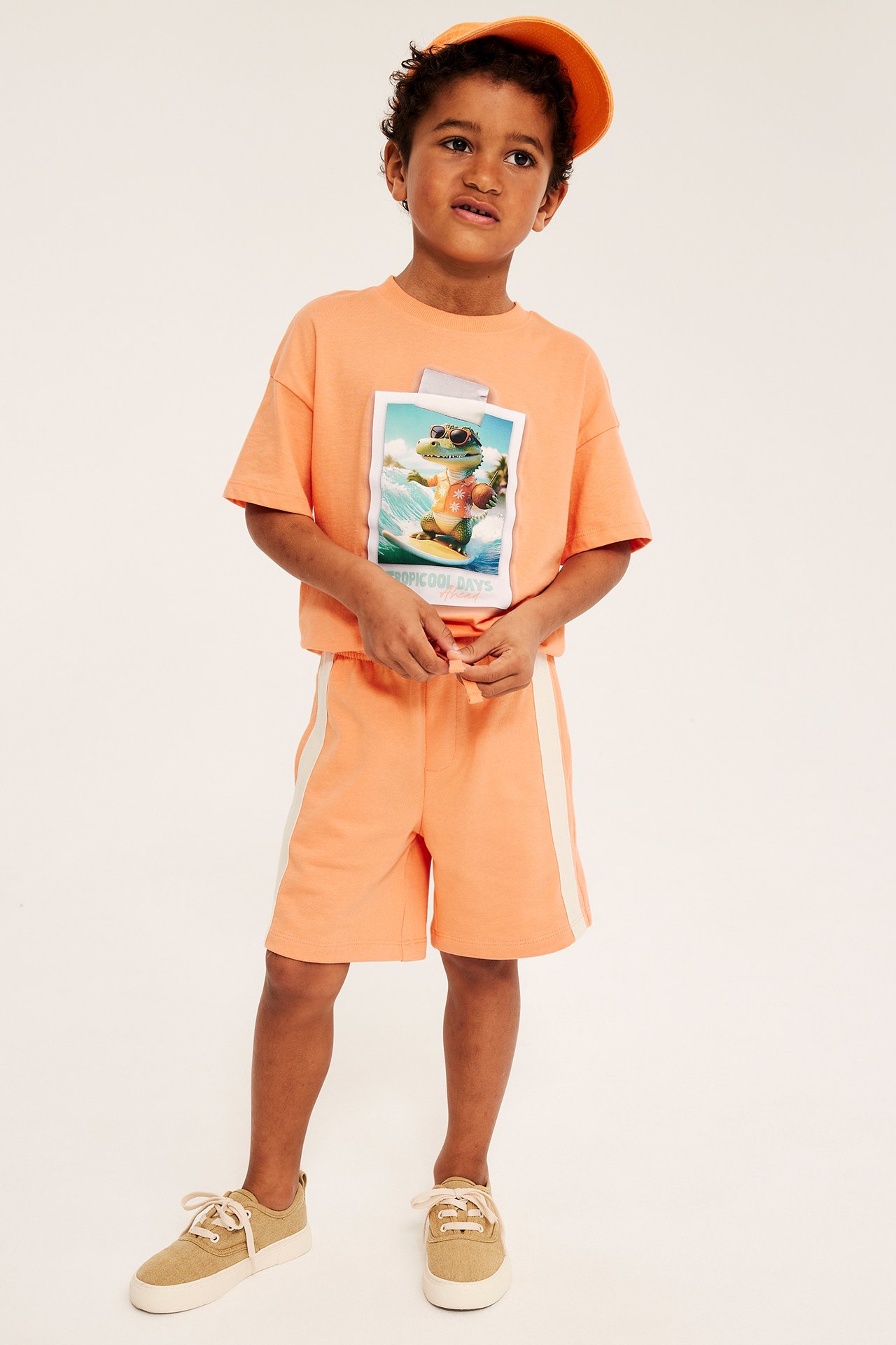 Edestä: Lapsi oransseissa collegeshortseissa valkoisilla raidoilla, kuvioitu t-paita, oranssi lippalakki, beiget tennarit.