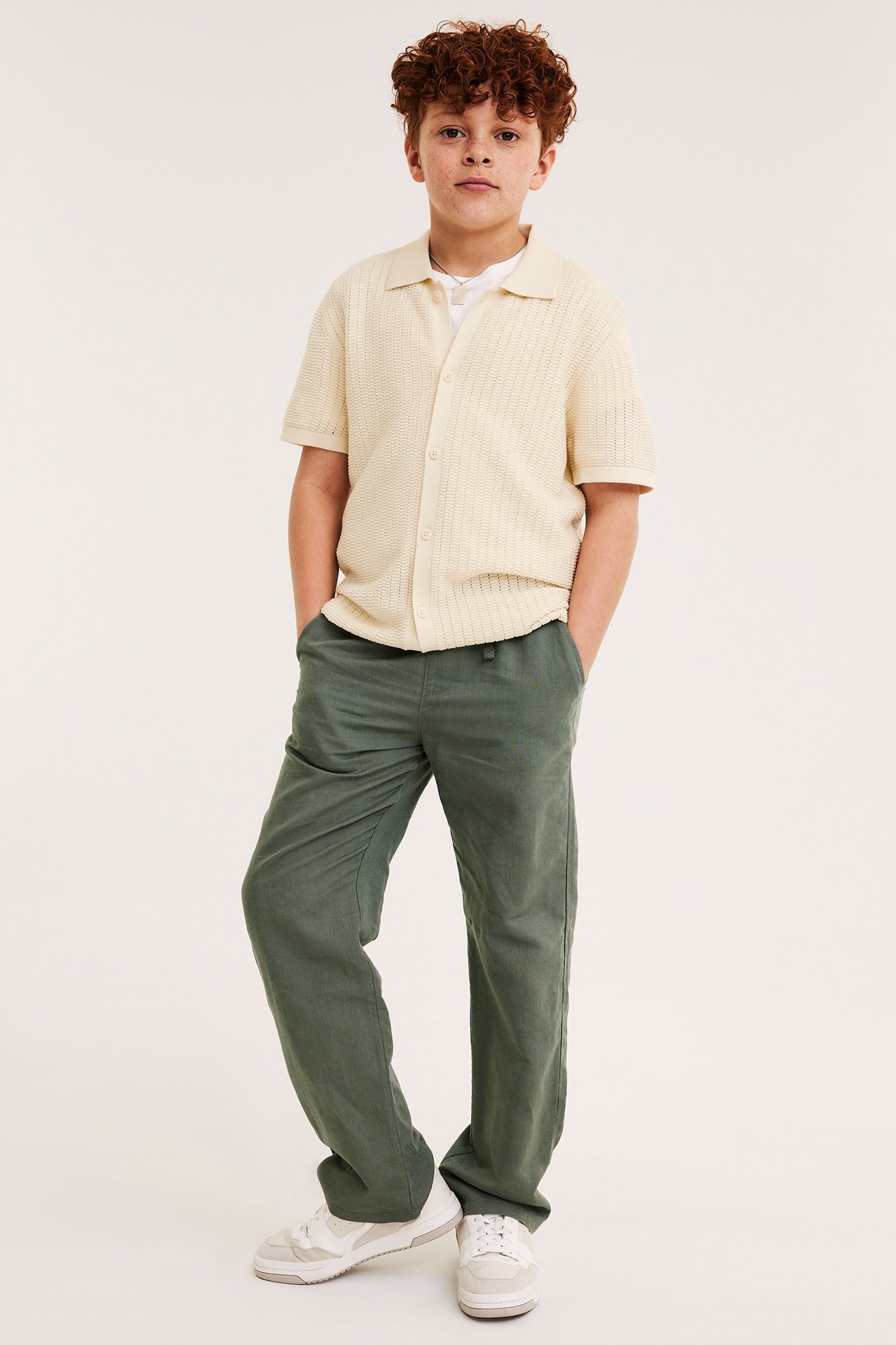 Edestä: poika vaaleanbeigessä neulotussa lyhythihaisessa paidassa valkoisen t-paidan ja vihreiden housujen kanssa.