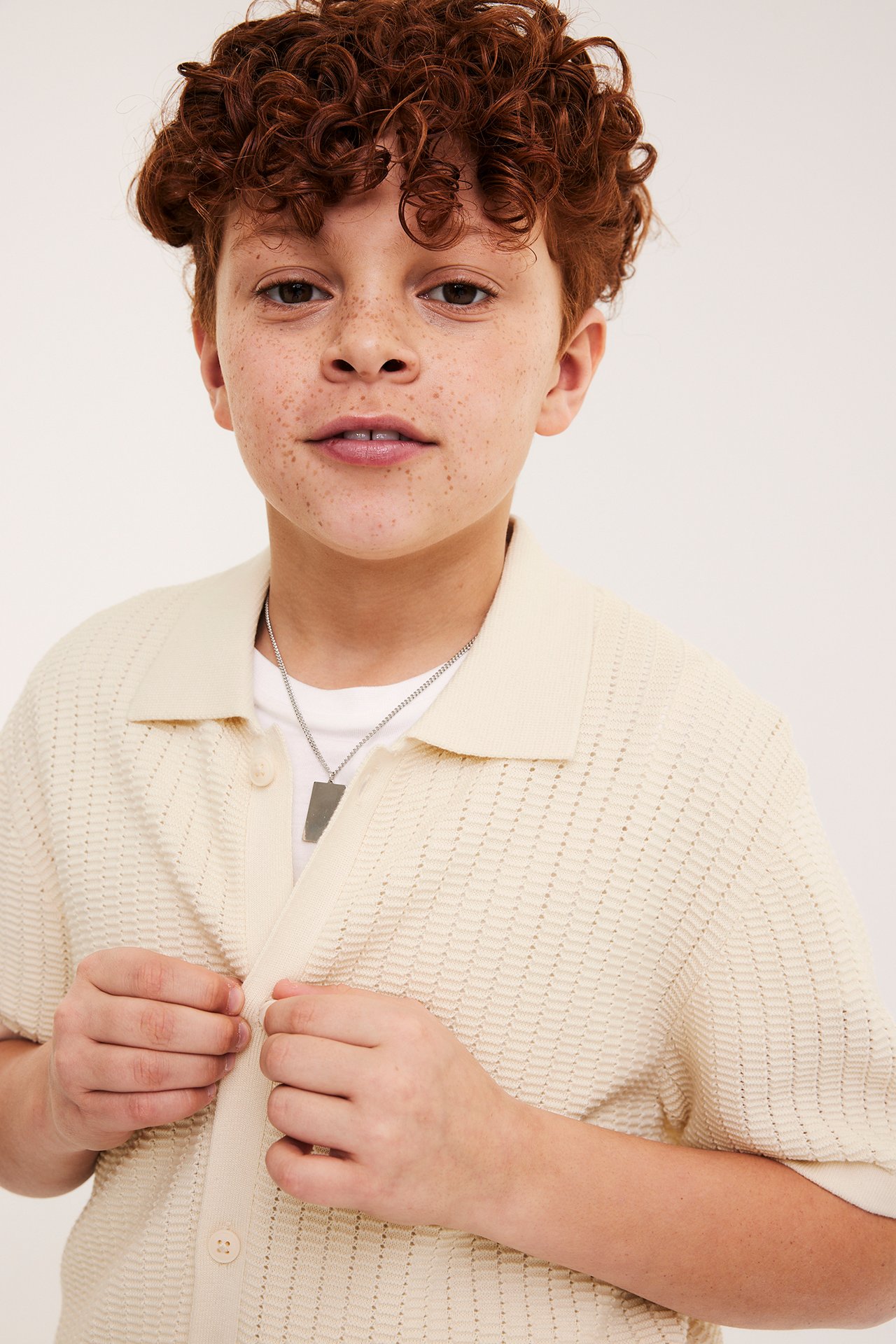 Barn knapper opp en lys beige kortermet jerseyskjorte over en hvit t-skjorte, med sølvkjede.