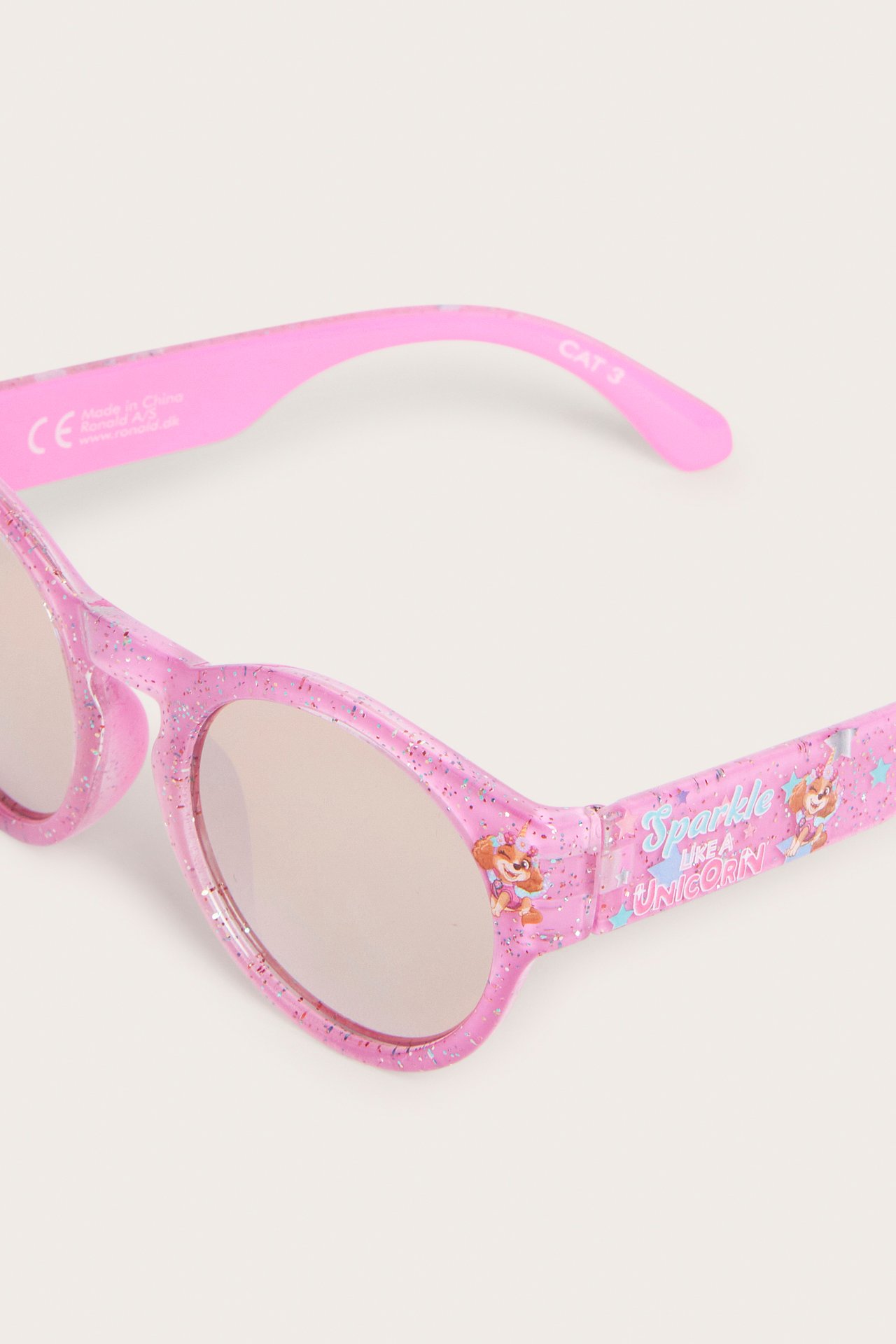Zbliżenie na różowe, brokatowe plastikowe okulary przeciwsłoneczne dla dzieci z nadrukiem Skye z Psiego Patrolu i napisem "Sparkle Like a Unicorn".