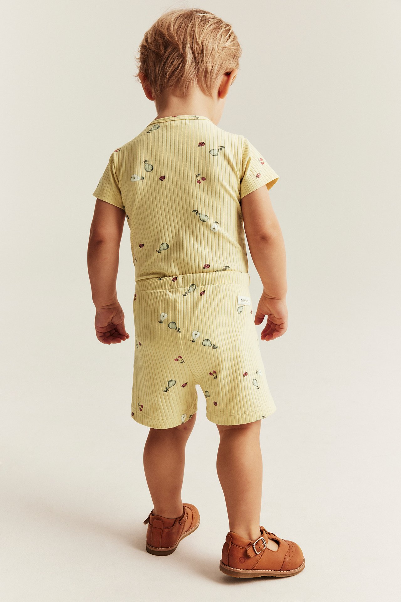 Bakfra: Barn i gult ribbet 2-delt sett med fruktmønster og brune spennesko.