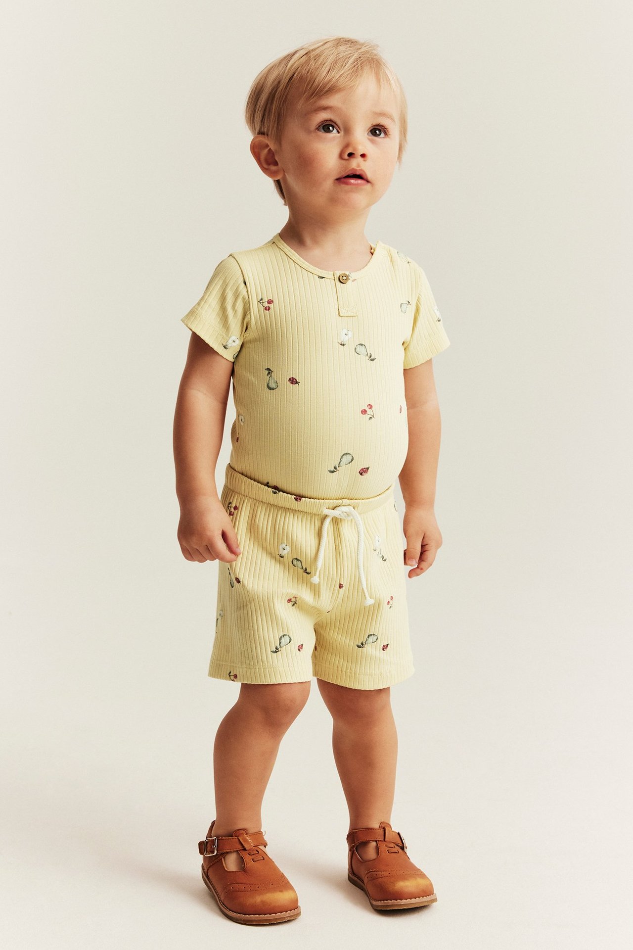 Forfra: en baby i gult ribbet kortermet body og shorts-sett med frukt- og marihønetrykk.