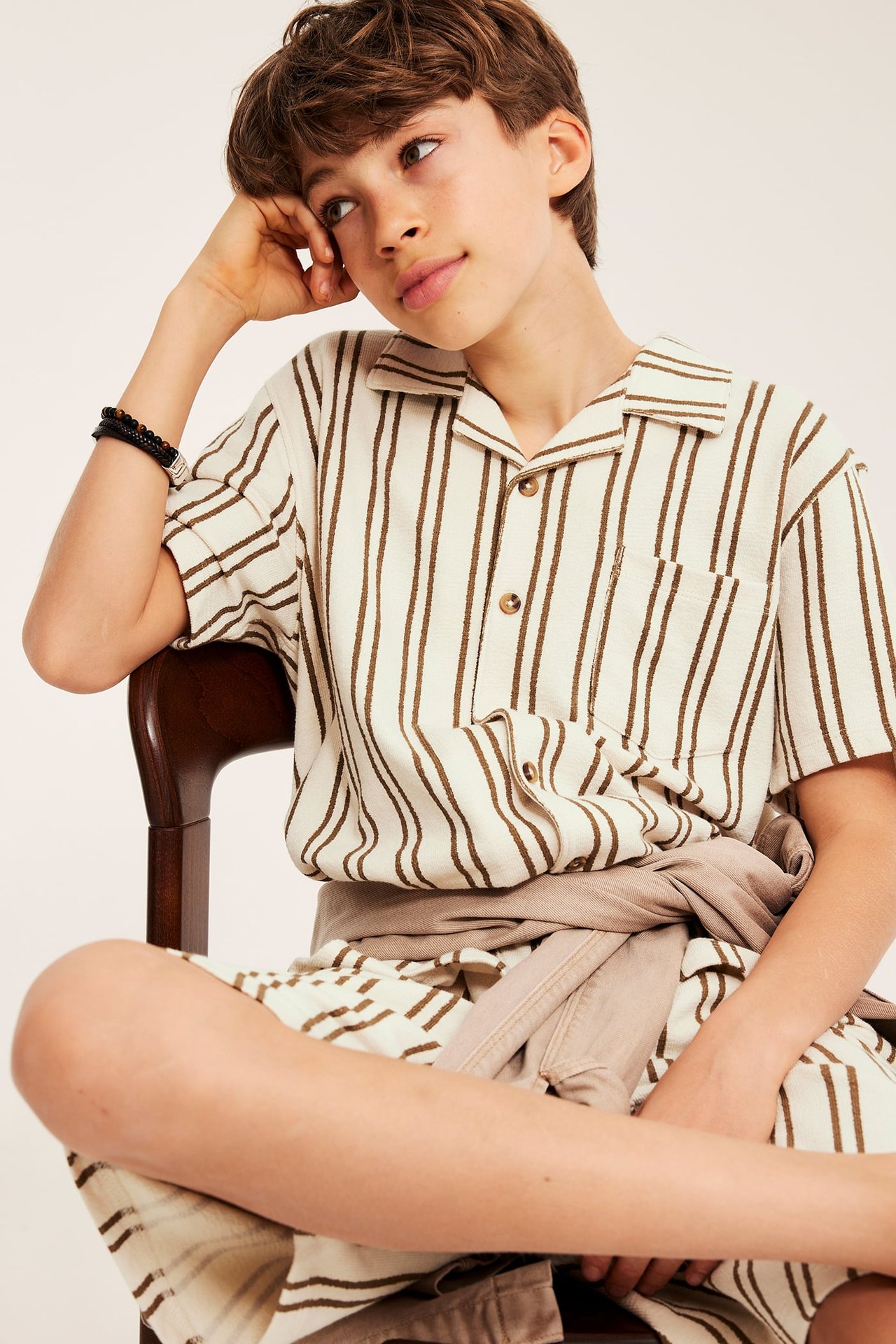 Framifrån, ett barn i en krämfärgad kortärmad randig skjorta med bruna ränder, matchande shorts och en beigetröja knuten.