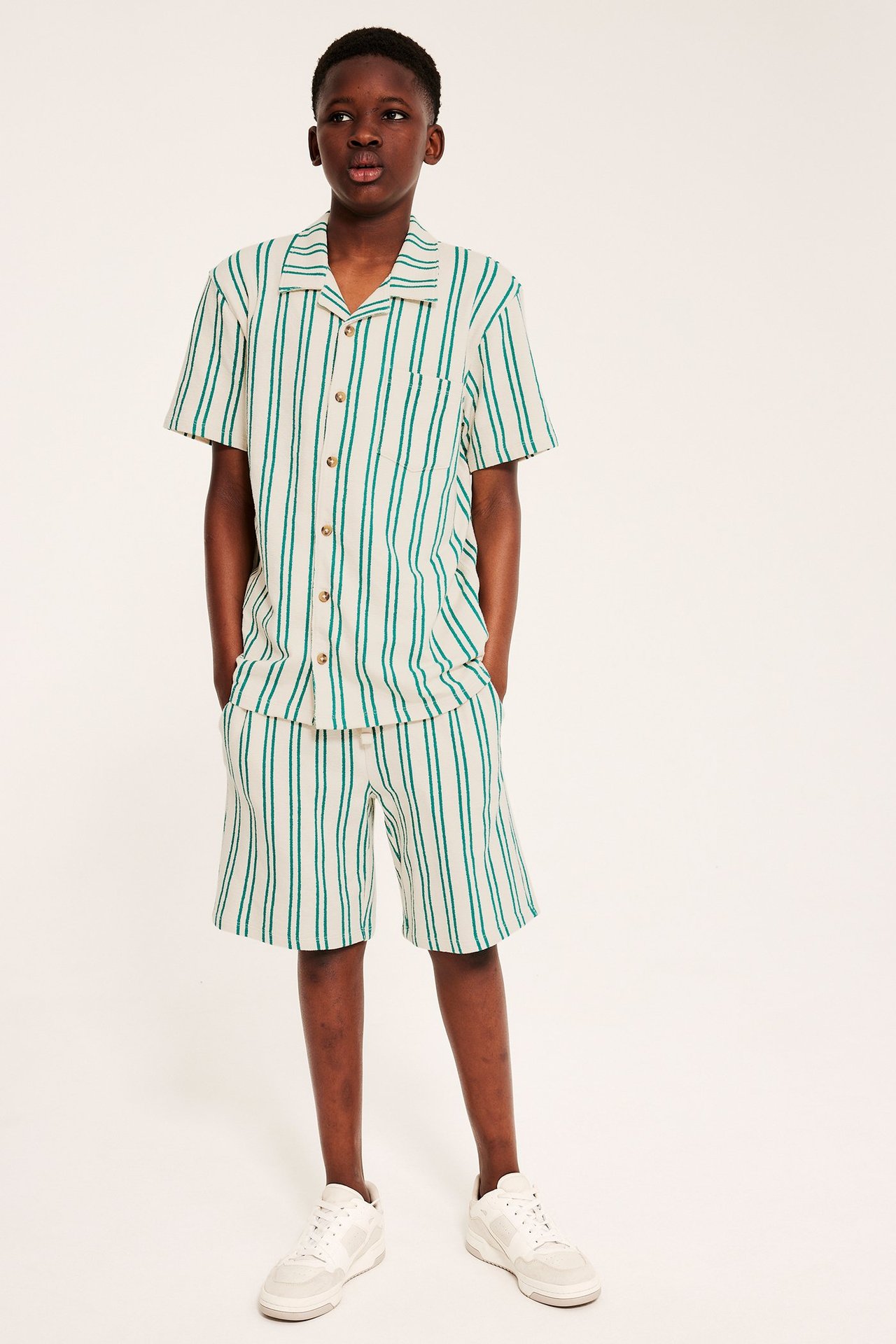 Frontvy av ett barn i grön- och benvitrandig kortärmad skjorta, matchande shorts och vita sneakers.