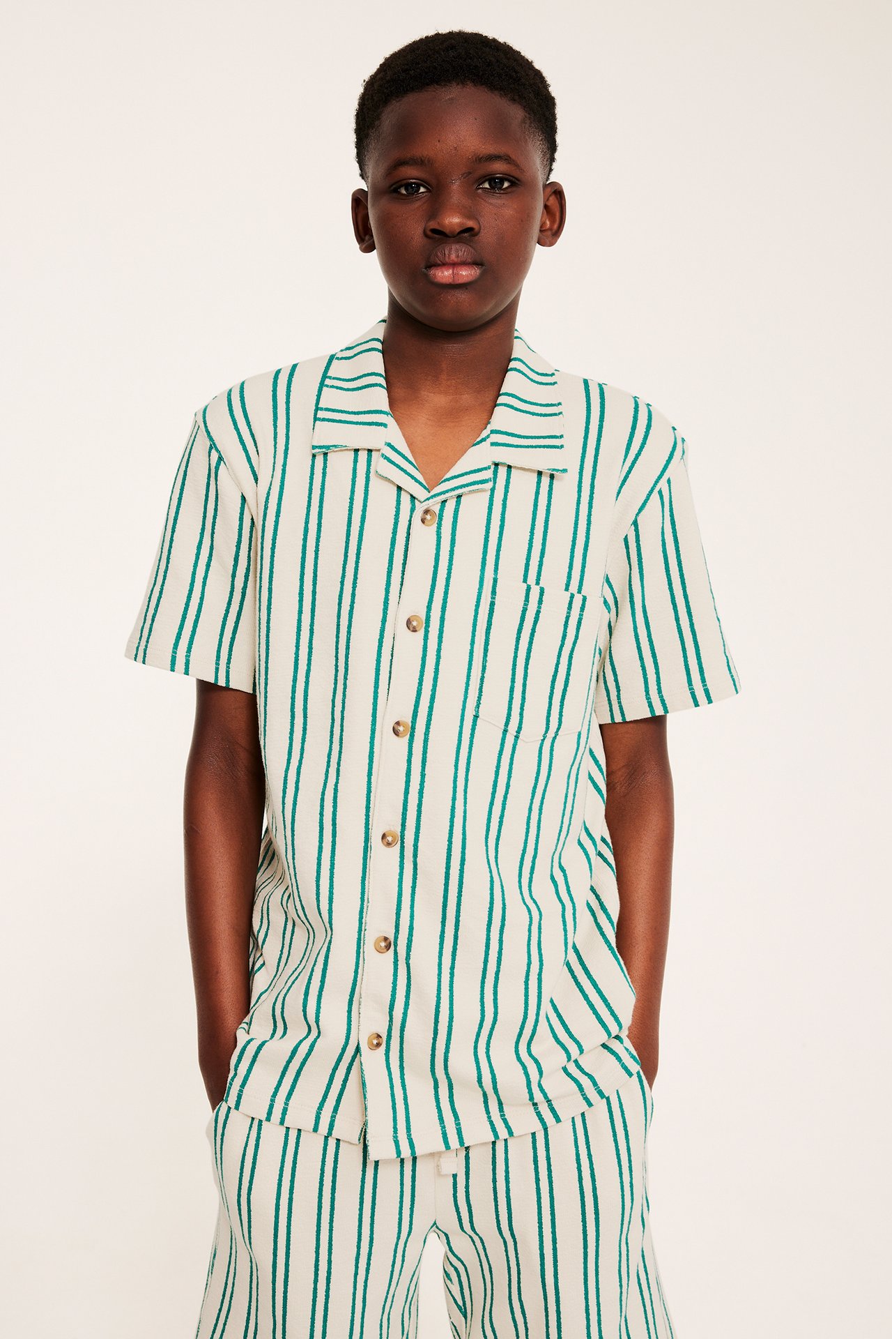 Front dziecka w kremowej, teksturowanej koszuli z krótkim rękawem w zielone paski i pasujących szortach.