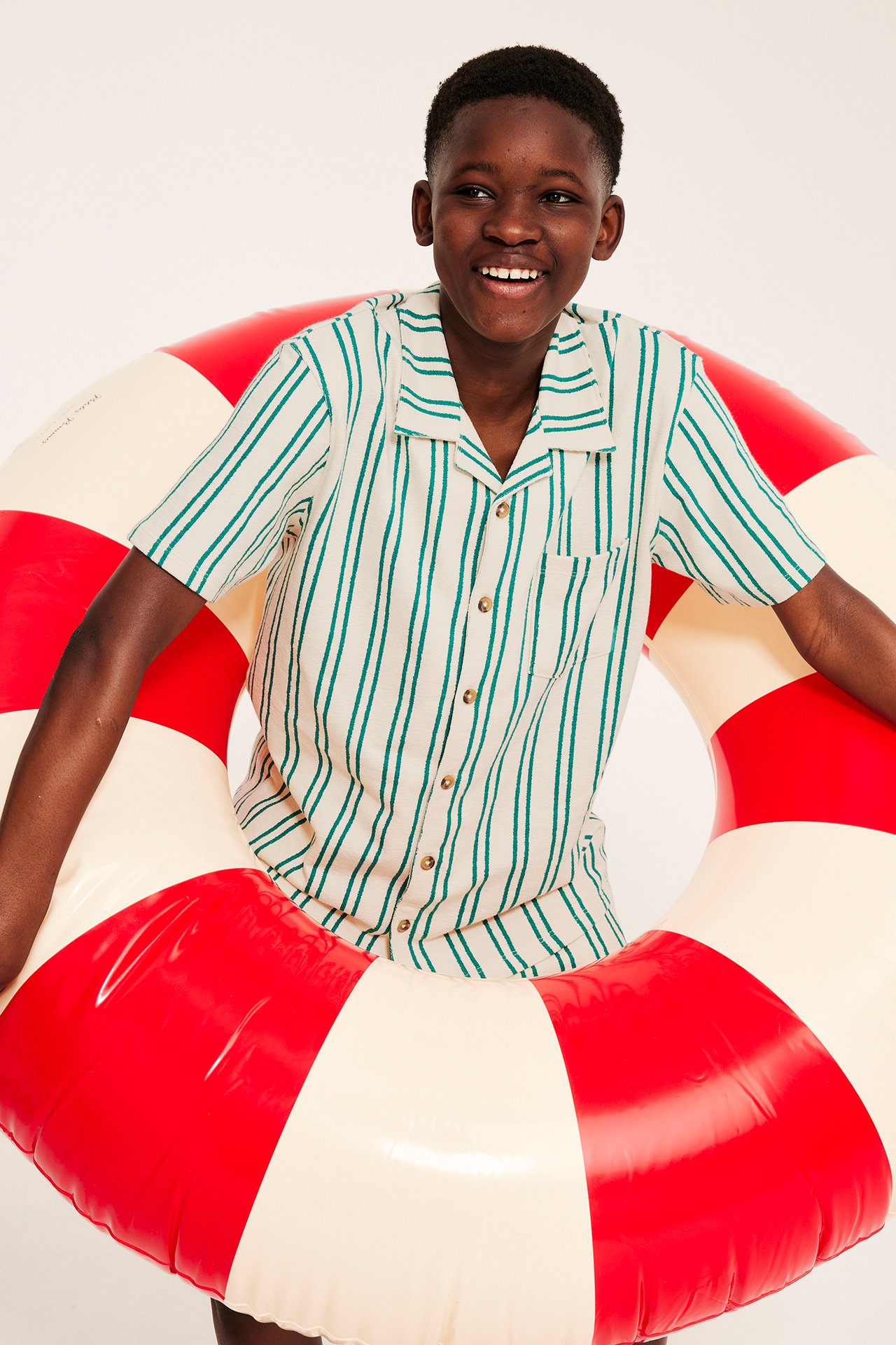 Uśmiechnięte dziecko w kremowej koszuli w zielone paski z krótkim rękawem, trzyma czerwono-białe kółko do pływania.