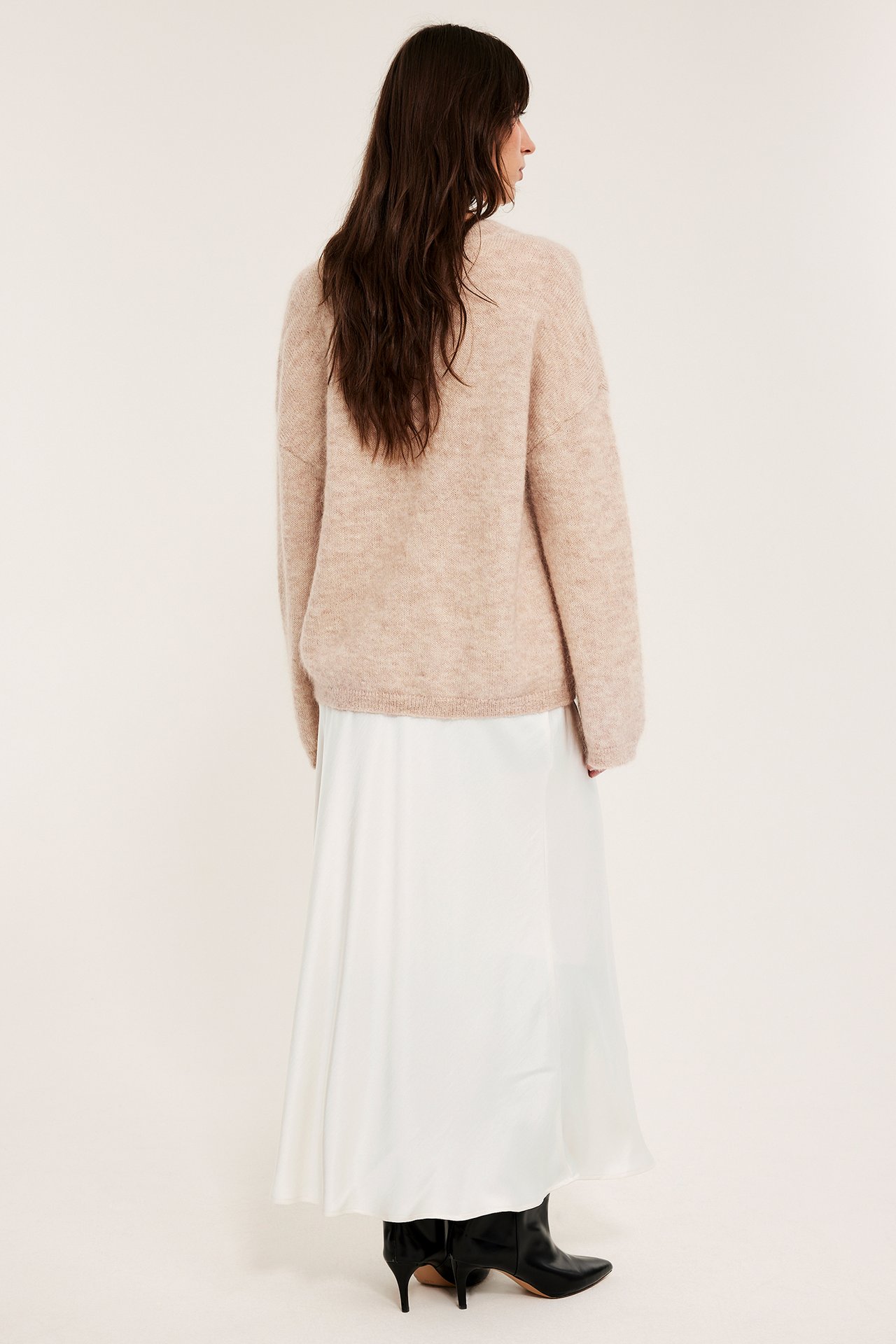 Widok z tyłu: kobieta w białej satynowej spódnicy, beżowym swetrze i czarnych botkach.