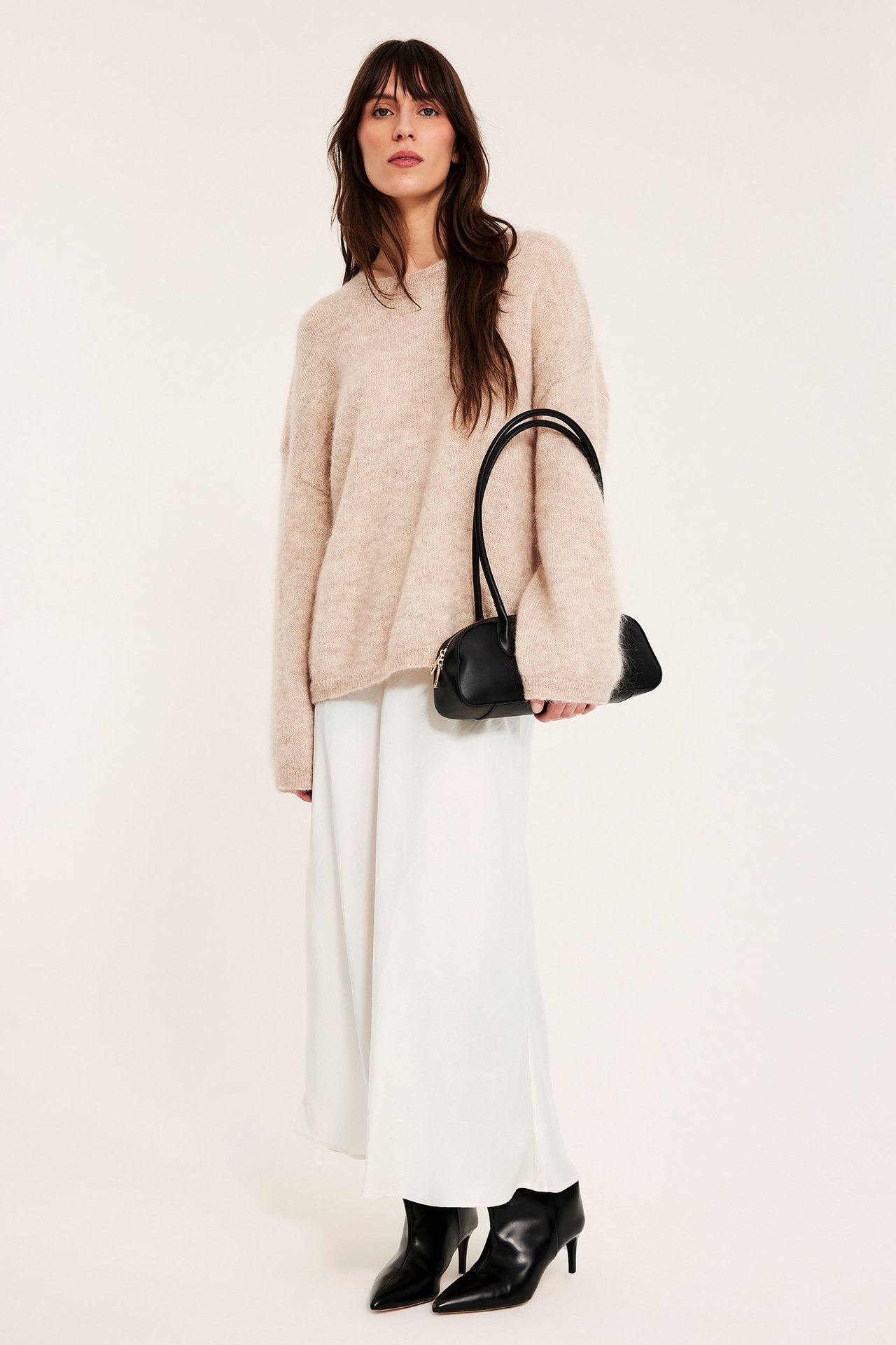 Frontvisning: kvinne i offwhite satengskjørt, beige genser, svarte ankelstøvler. Holder svart veske.