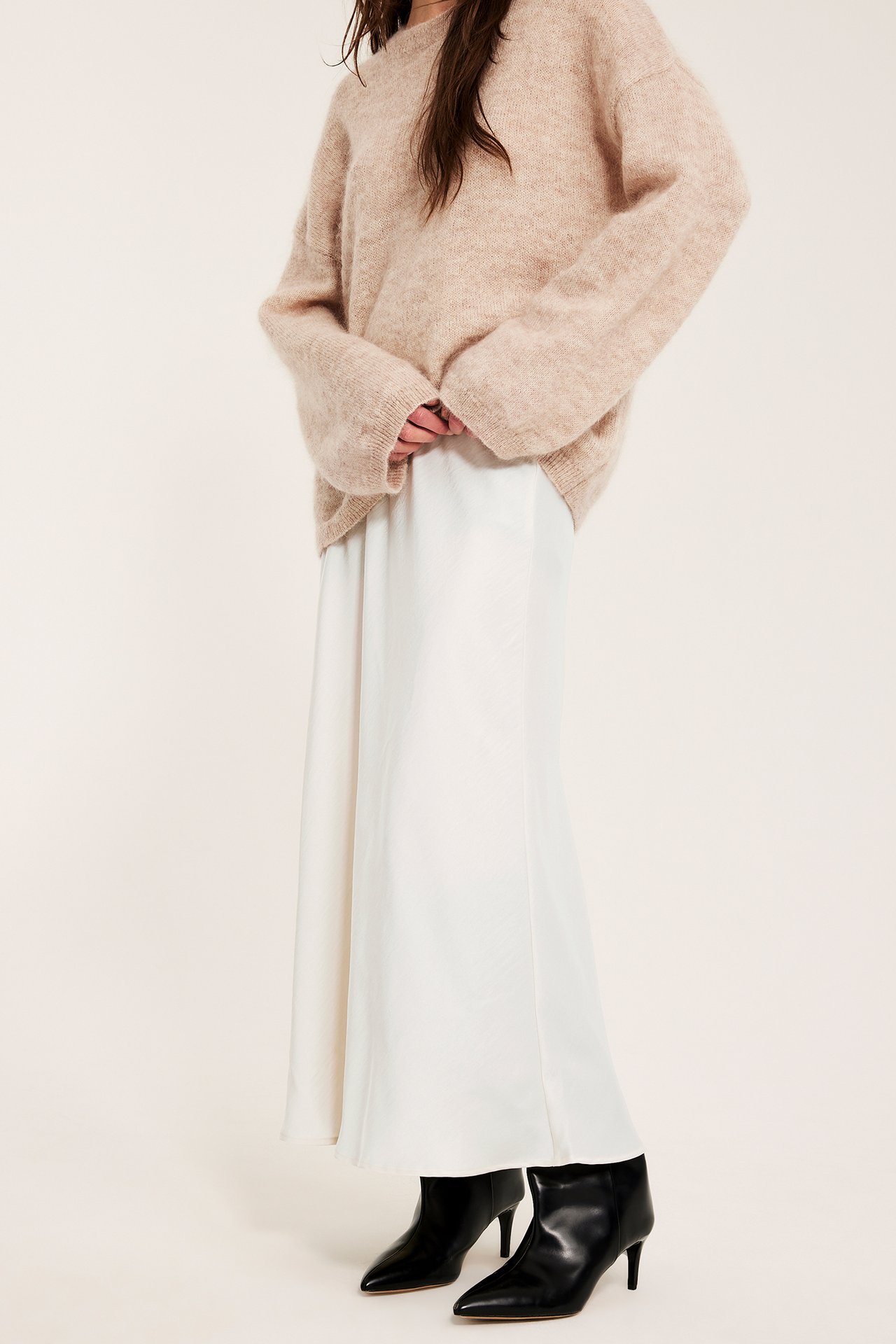 Widok z przodu kobiety w spódnicy satynowej rozkloszowanej off-white, beżowym swetrze i czarnych botkach.