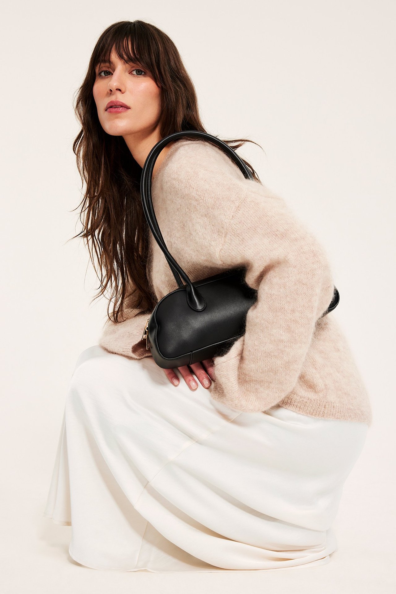 Przód: kobieta w spódnicy z rozkloszowanej satyny w kolorze złamanej bieli, beżowym swetrze, trzyma czarną torebkę.