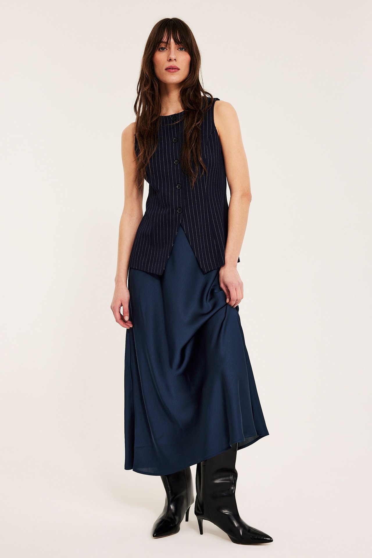 Frontvy: kvinna i mörkblå utsvängd satinkjol, marinblå nålränd ärmlös topp och svarta stövlar.