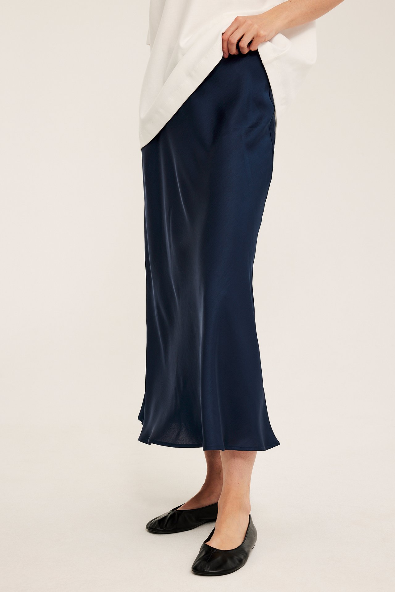 Sidovy: kvinna i mörkblå klockad satinkjol av polyester, vit topp och svarta platta skor.