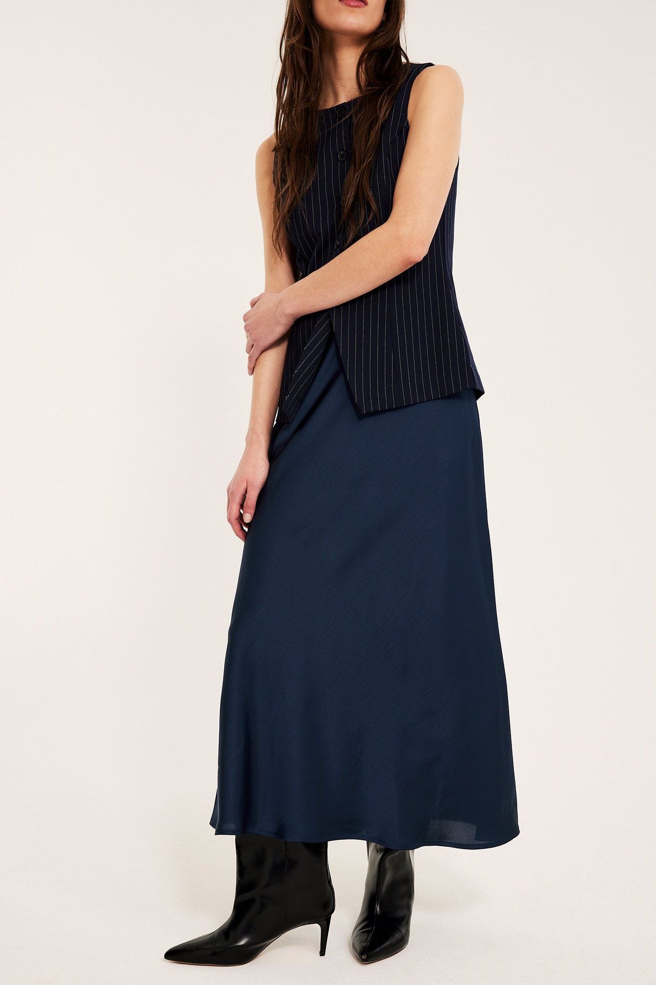 Widok z przodu: Kobieta w ciemnoniebieskiej rozkloszowanej spódnicy satynowej, prążkowanym topie bez rękawów i czarnych botkach.