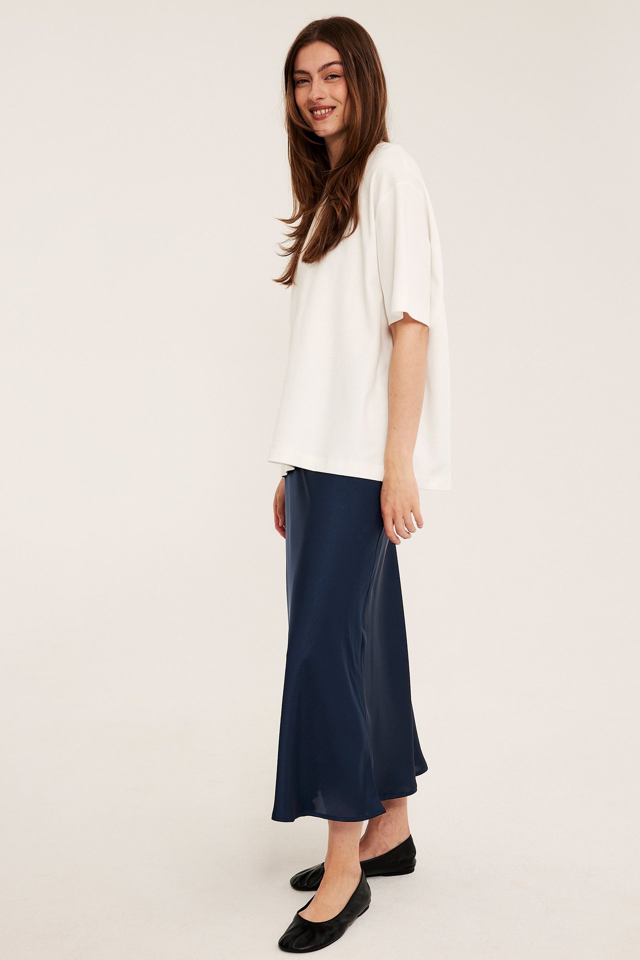Sidovy: Kvinna bär mörkblå klockad satinkjol, en off-white t-shirt och svarta ballerinaskor.