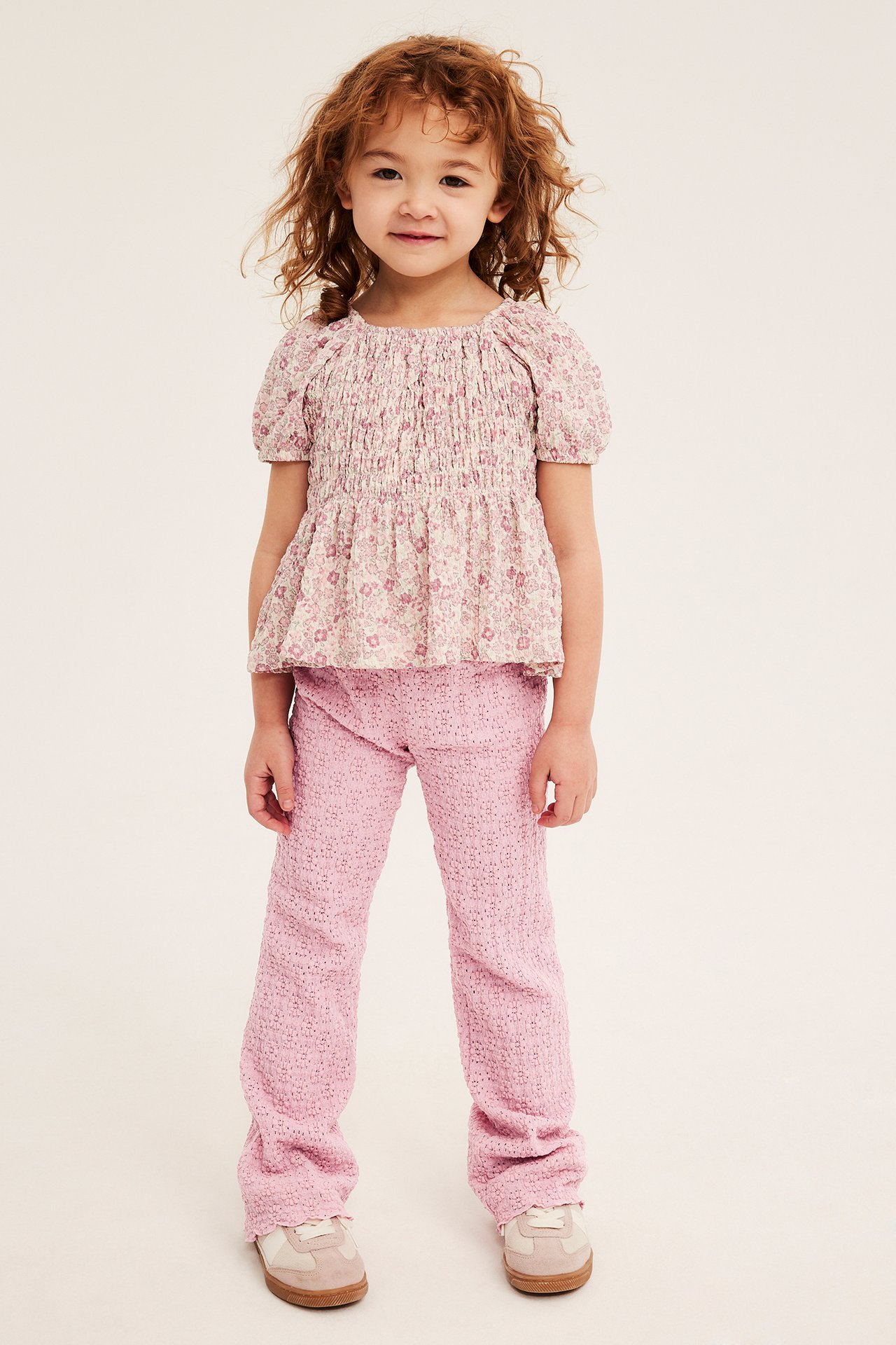Frontvisning av et barn i lyserosa utsvingte blondeleggings, en blomstret smocket topp og joggesko.