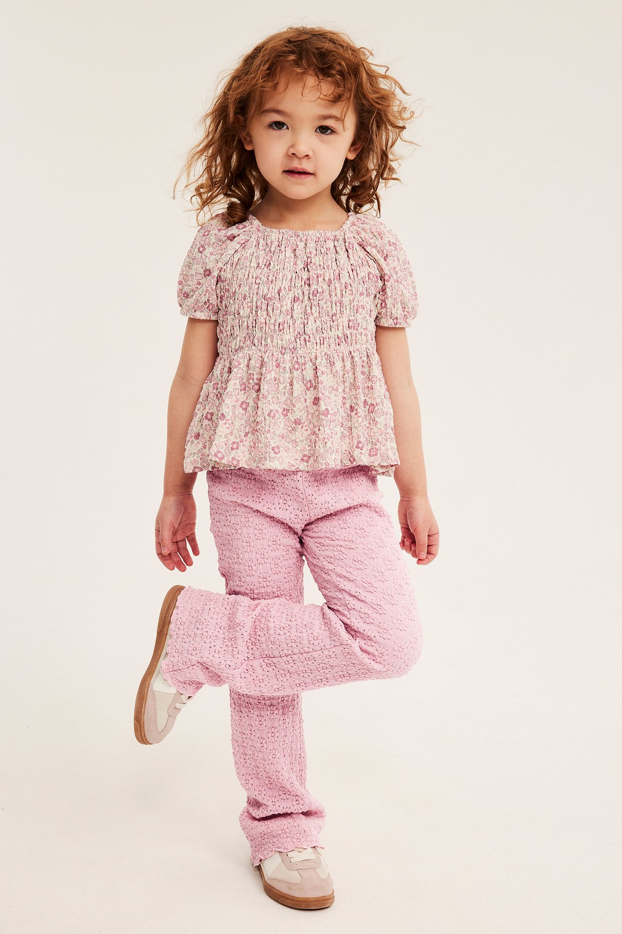 Barn i lyserosa utsvingte blondeleggings, blomstret smockbluse og lyse sneakers, forfra.