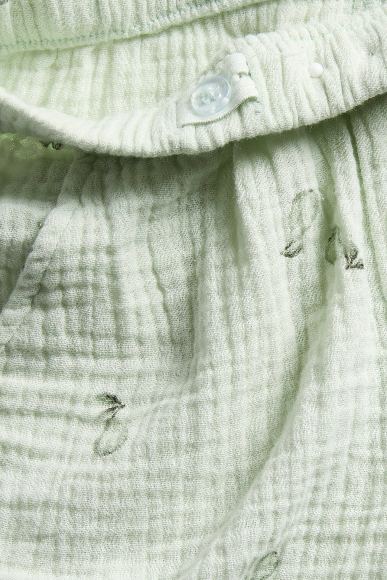 Nærbilde: lysegrønne bomullsmuslinshorts med pæremønster og dekorativ knapp i strikk i midjen.