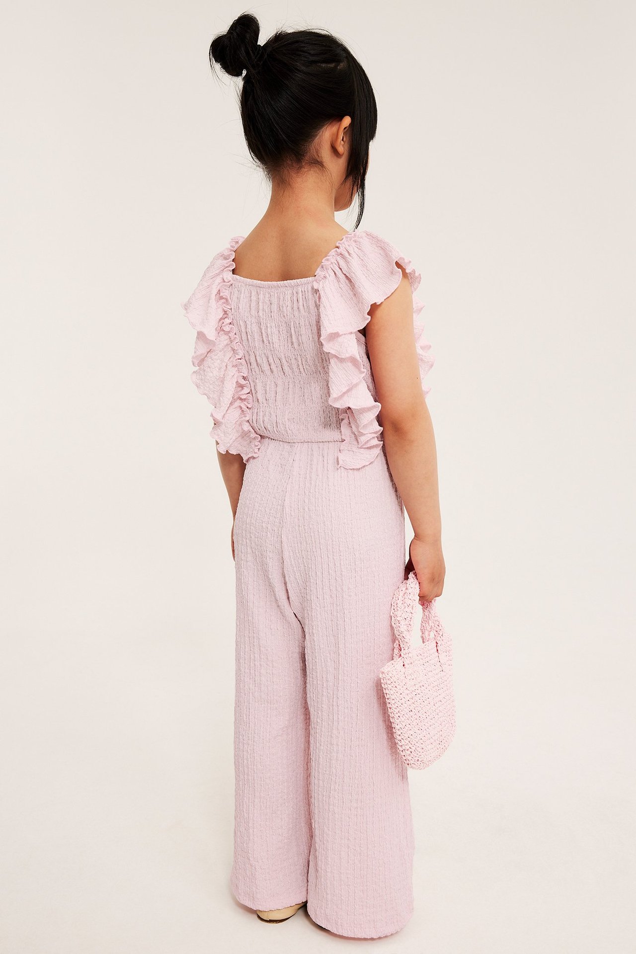 Dziecko w jasnoróżowym kombinezonie z dżerseju, falbanki, szerokie nogawki, trzymające różową dzierganą torebkę.