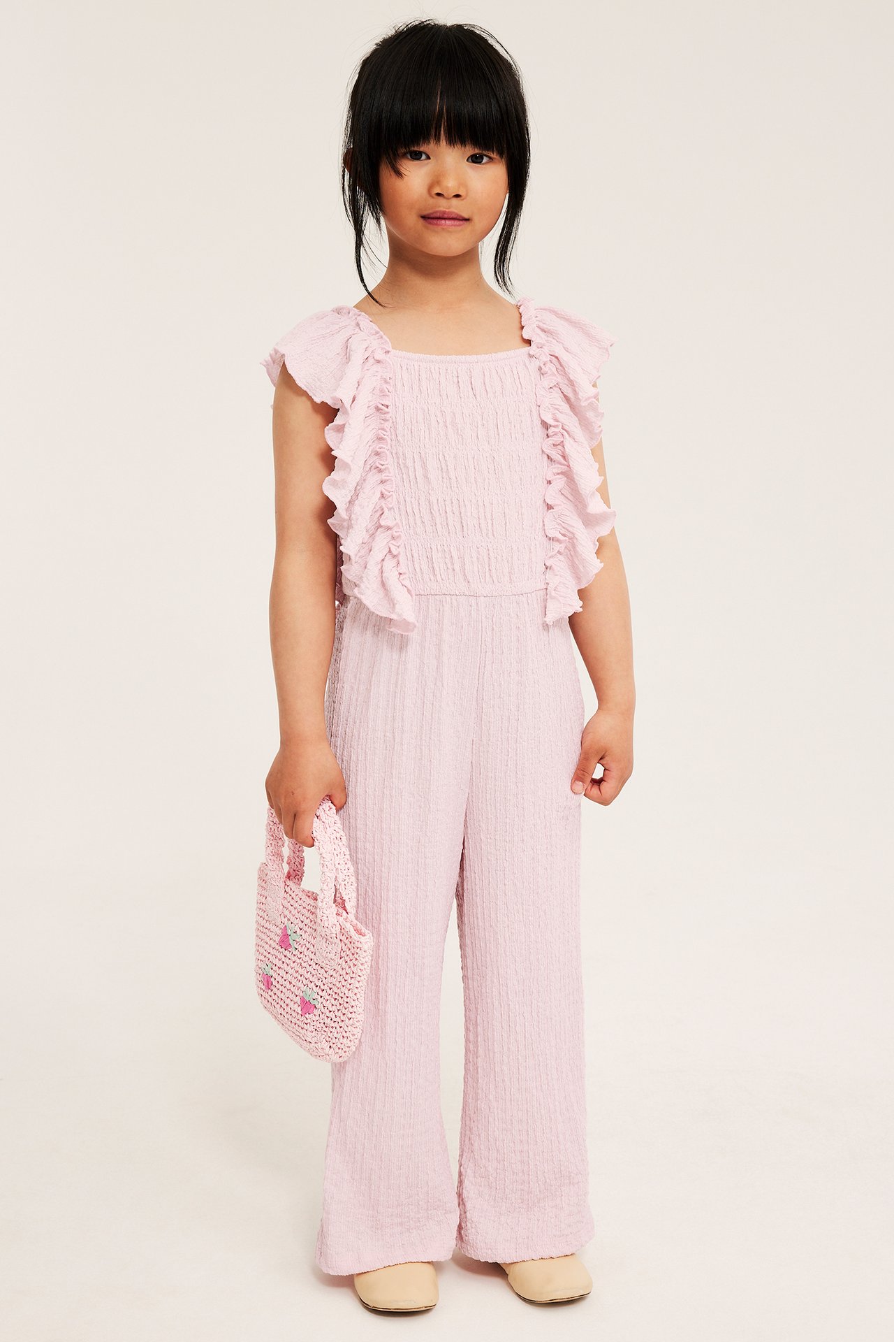 Edestäpäin lapsi vaaleanpunaisessa röyhelöisessä jerseyhaalarissa, pitää vaaleanpunaista mansikkalaukkua.