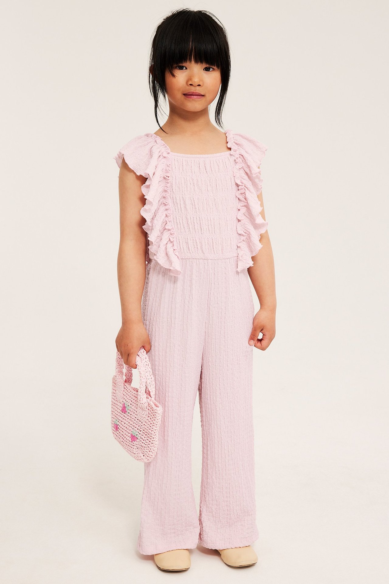 Edestäpäin lapsi vaaleanpunaisessa röyhelöisessä jerseyhaalarissa, pitää vaaleanpunaista mansikkalaukkua.